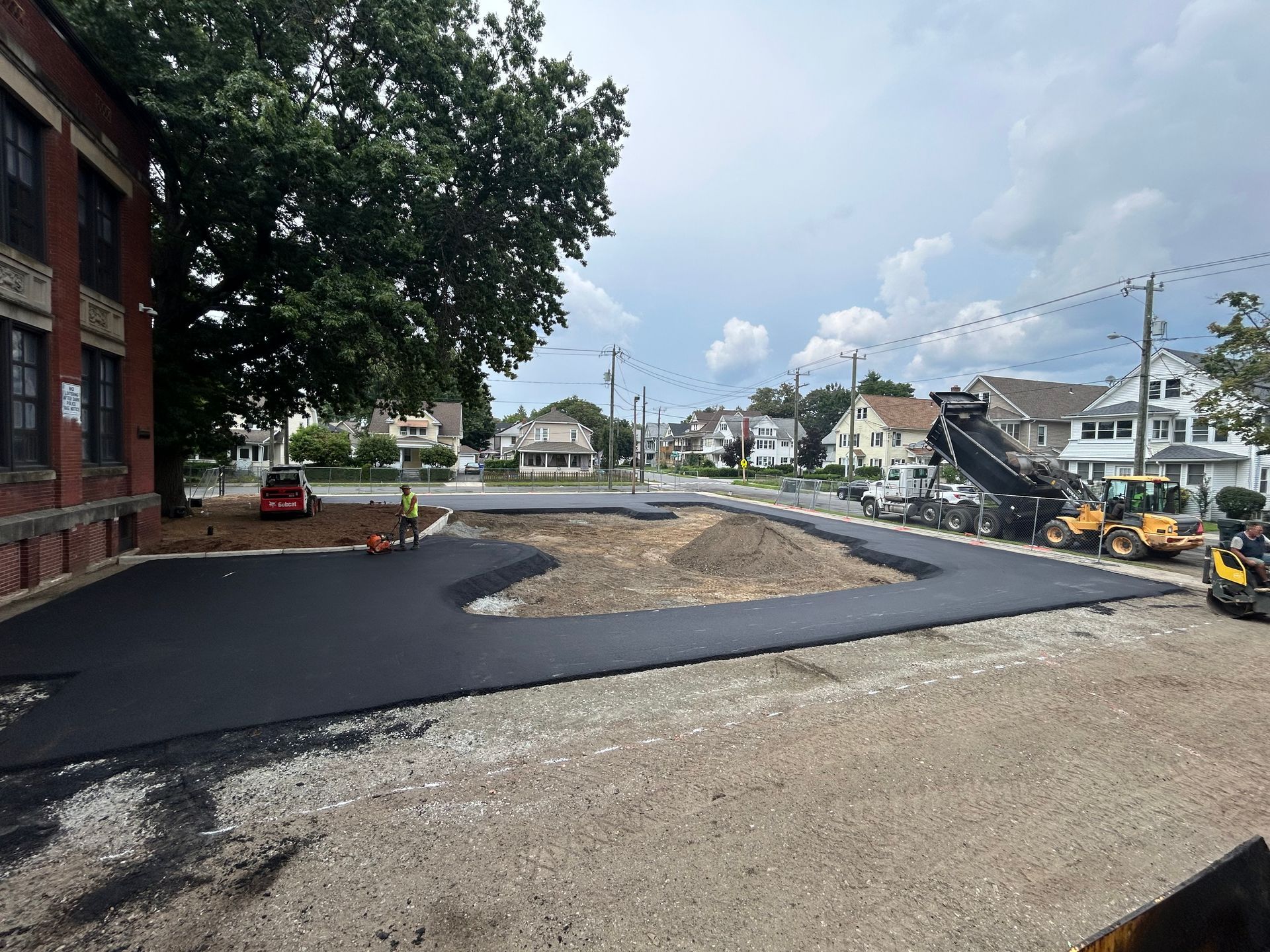 Construction crew paving a parking lot with asphalt next to a brick building and residential houses on a sunny day.