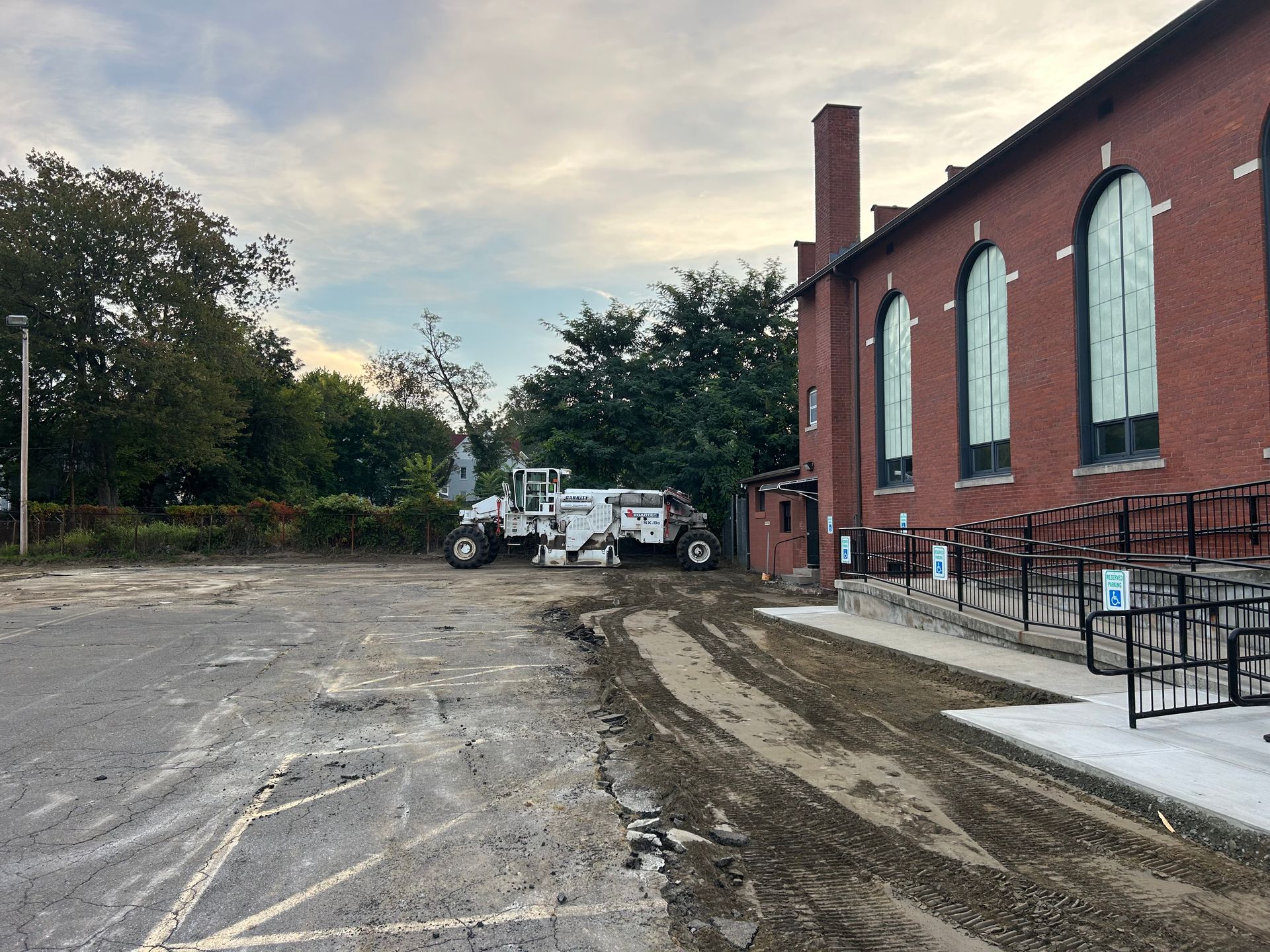 A white road construction vehicle parked next to a red brick building with arched windows, beside a gravel lot.