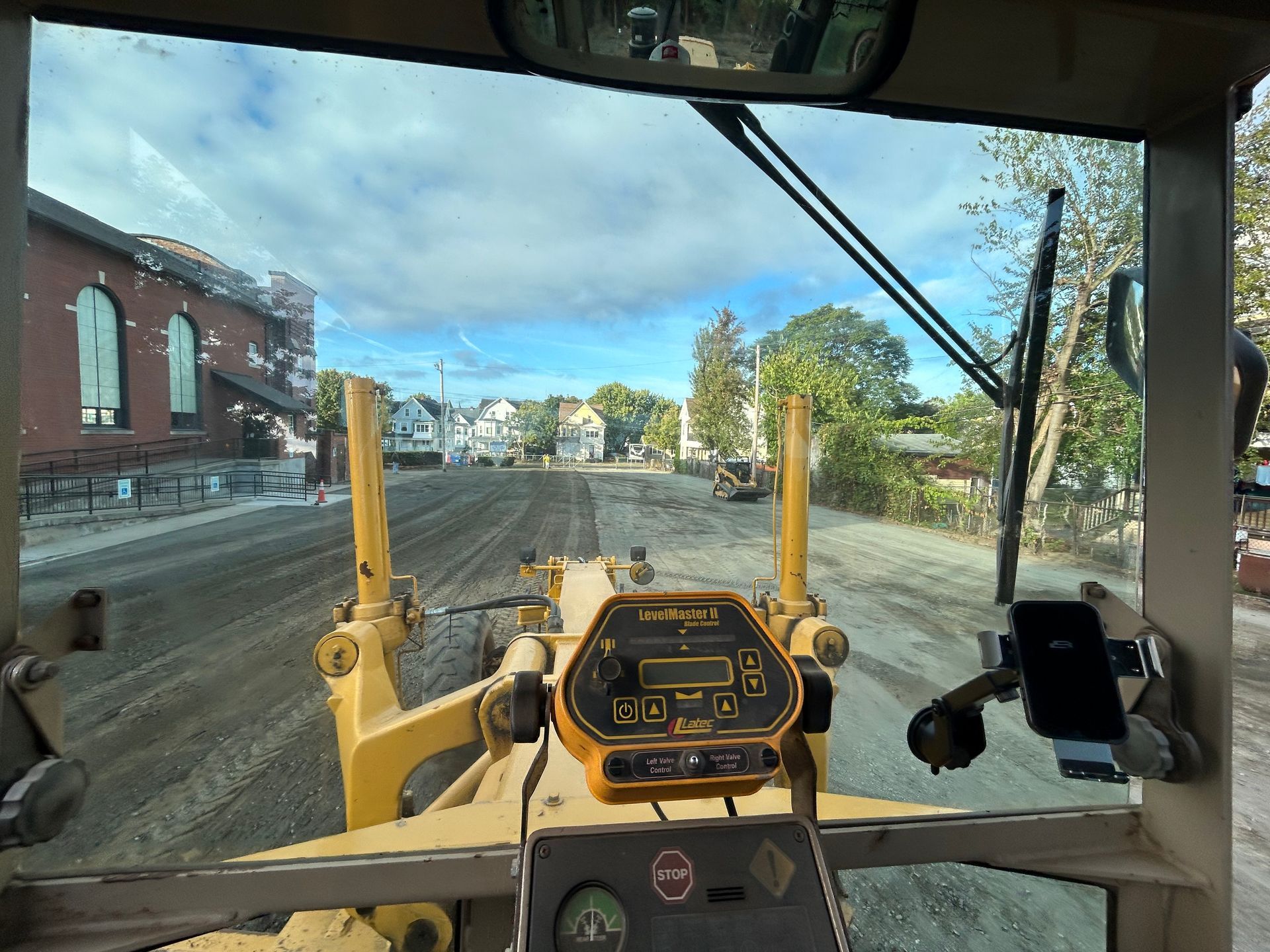 View from the cab of a yellow motor grader on a dirt road, facing houses under a blue sky with clouds.