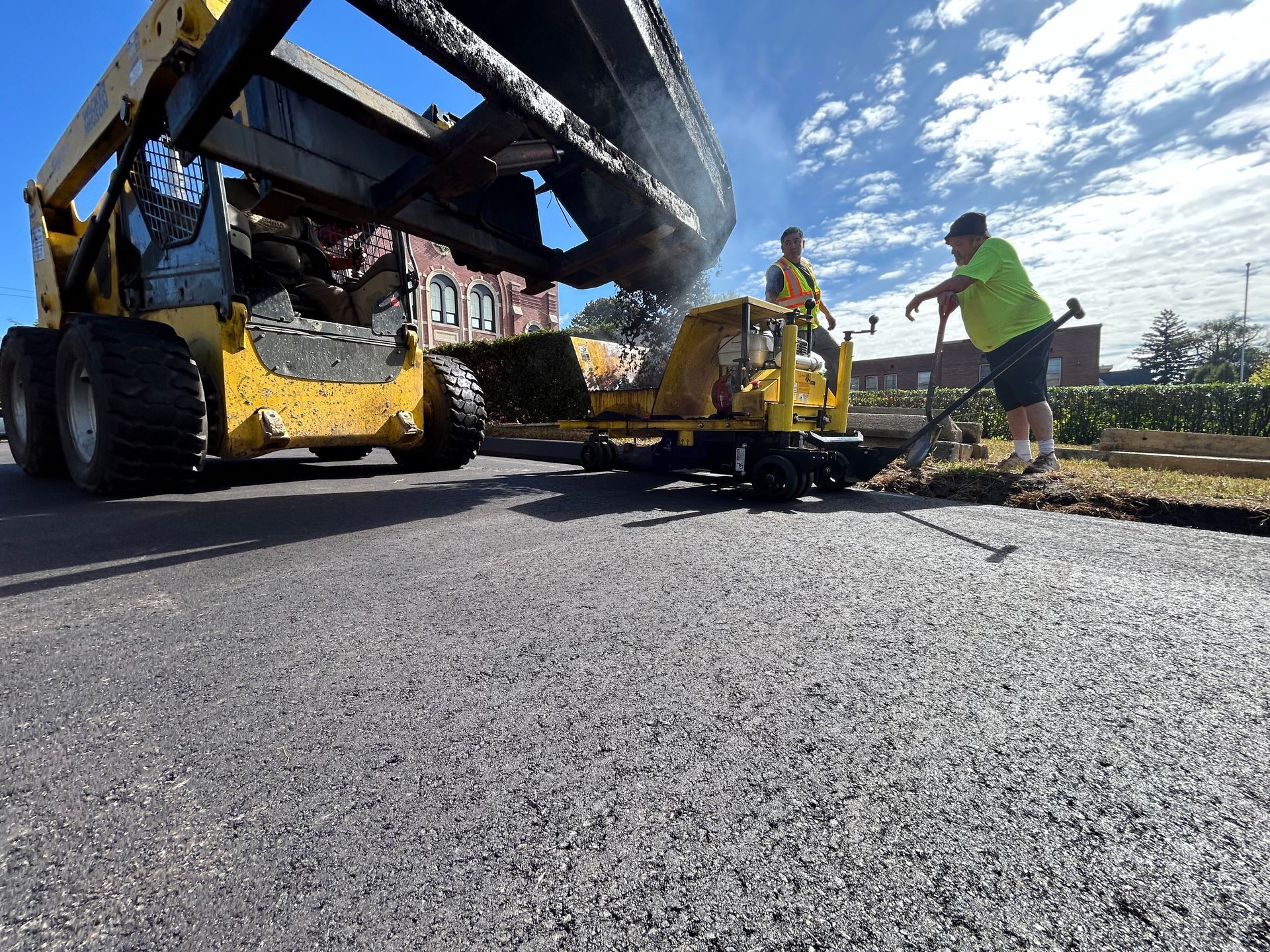 Two workers in safety vests use a yellow skid-steer loader to pour asphalt on a sunny street.