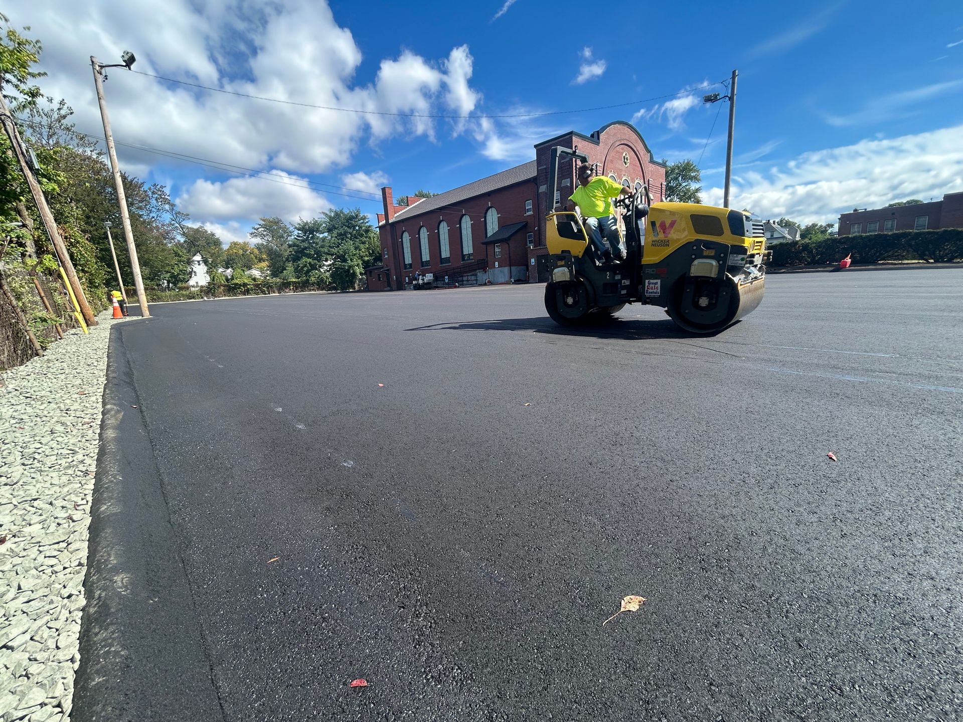 A worker in a high-visibility yellow vest operates a yellow asphalt roller on a newly paved parking lot beside a building.