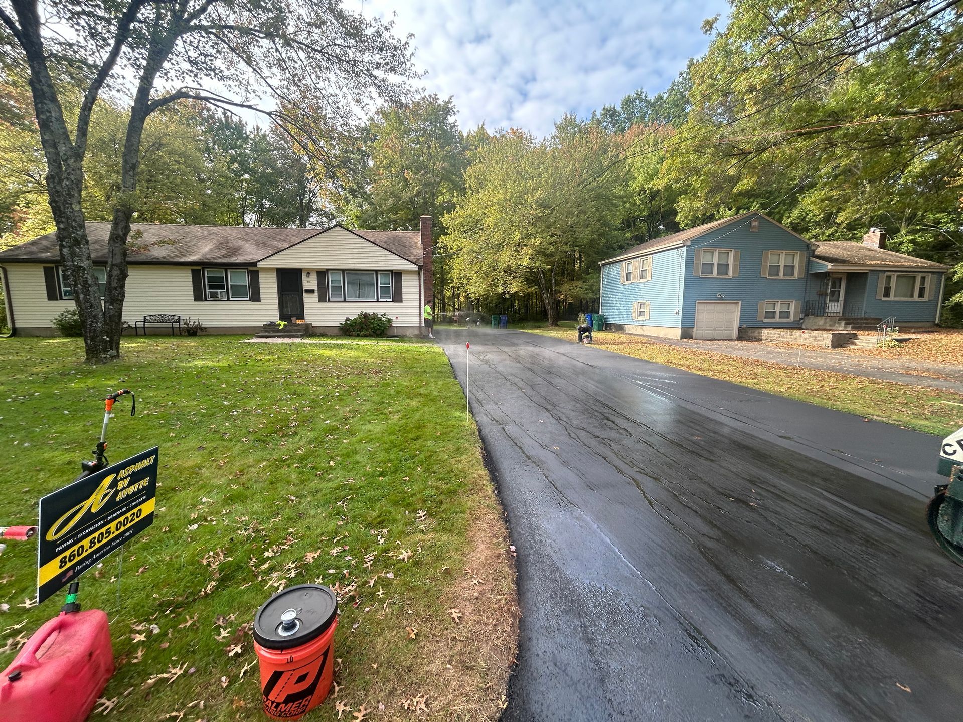A freshly paved asphalt driveway leading between two suburban houses with lawns and surrounding trees under a cloudy sky.