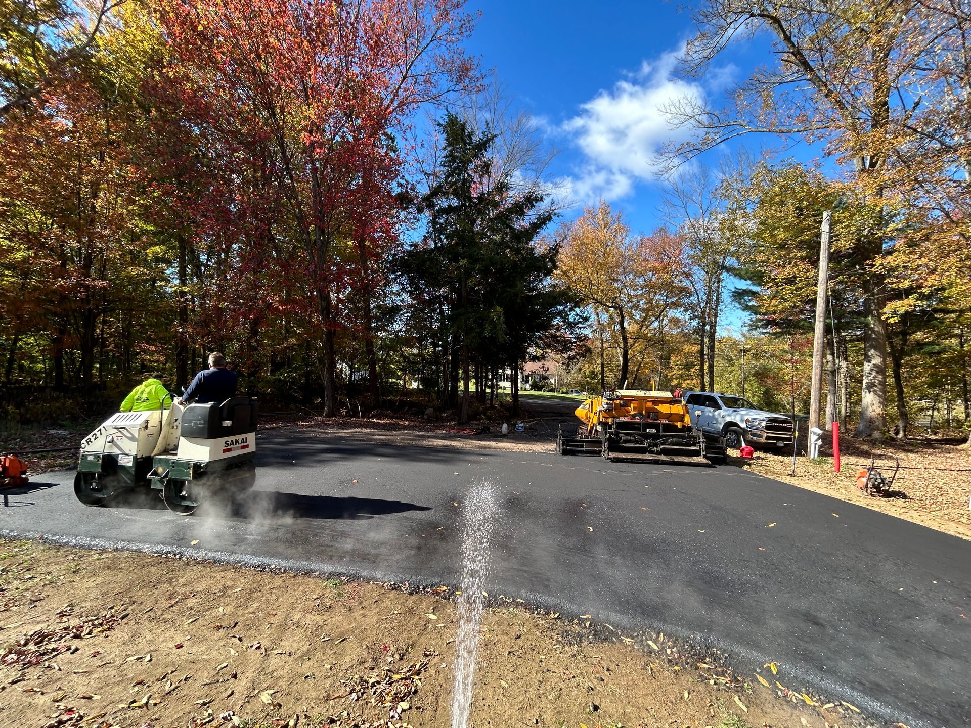A worker operates a white asphalt roller to smooth a newly paved driveway, with trees in autumn foliage in the background.