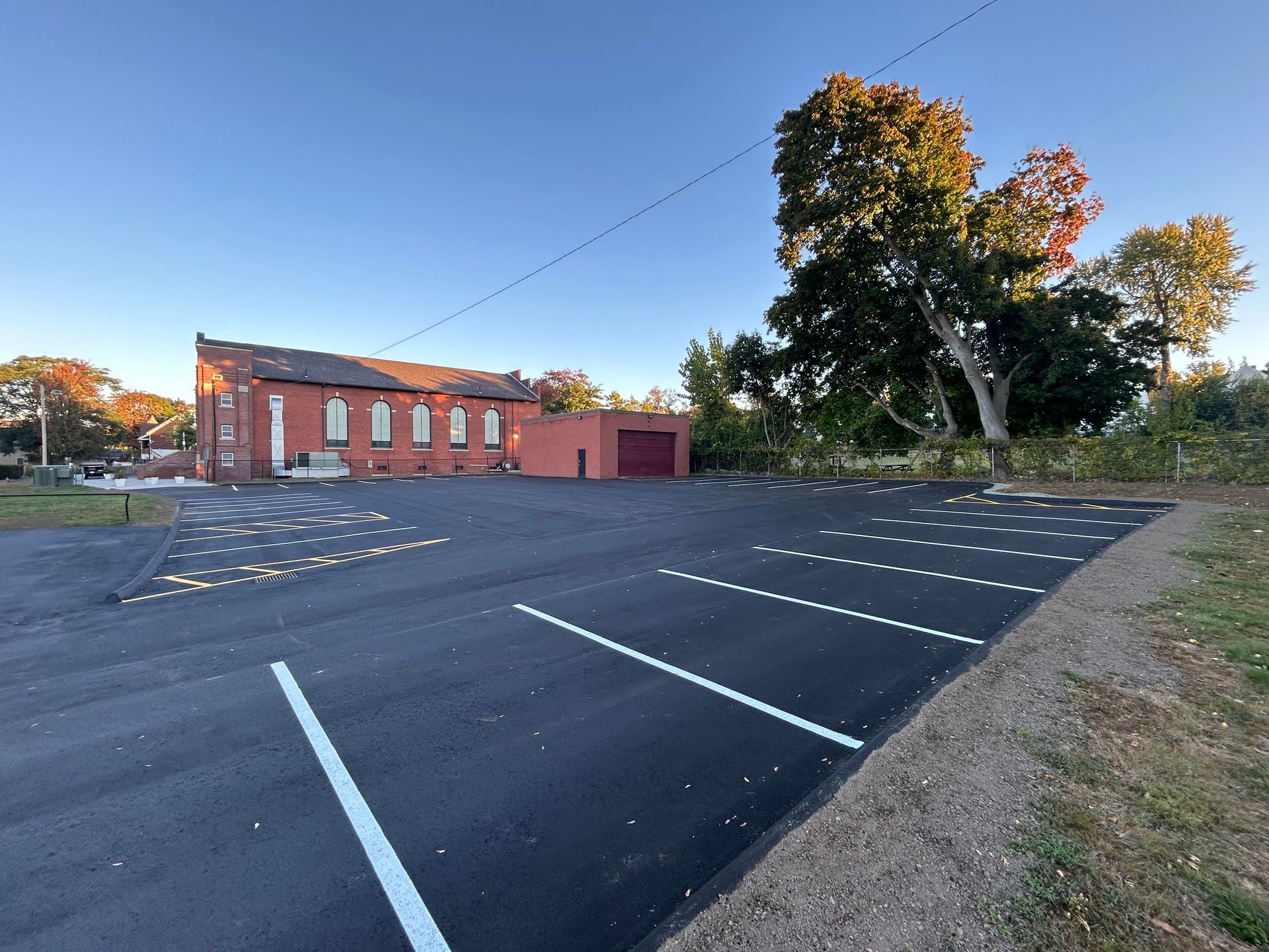 A freshly paved asphalt parking lot with white painted stalls next to a brick building and a large tree under a blue sky.