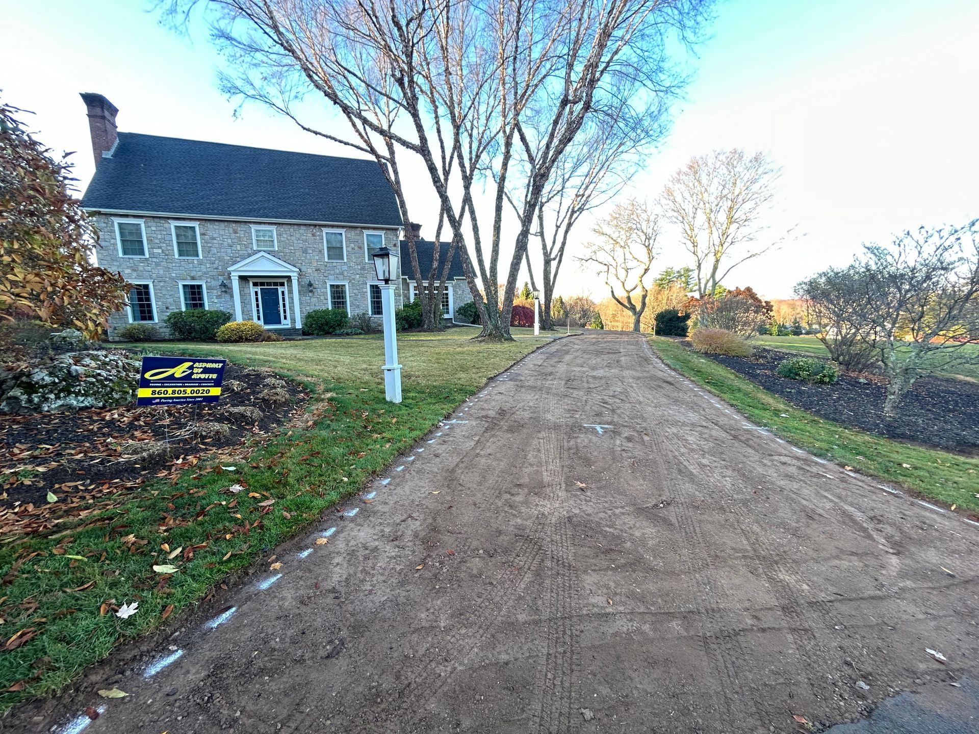 A stone colonial house with a dark roof stands behind a freshly graded, unpaved dirt driveway.