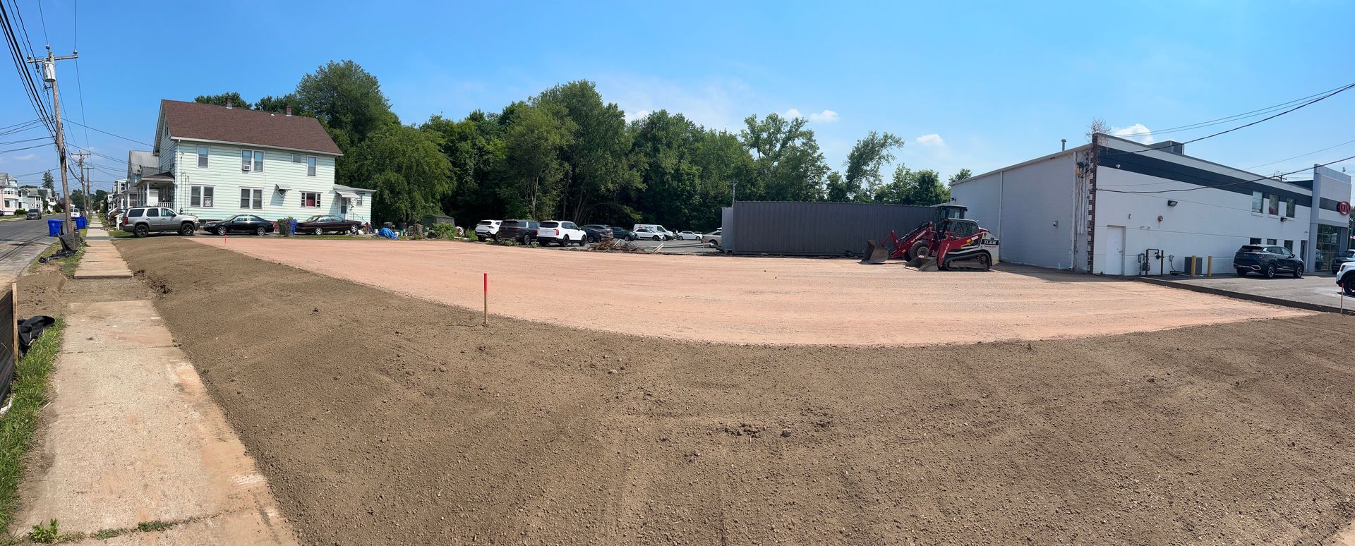 A flat, cleared lot of light-colored dirt bordered by a sidewalk, a white building, trees, and a house.