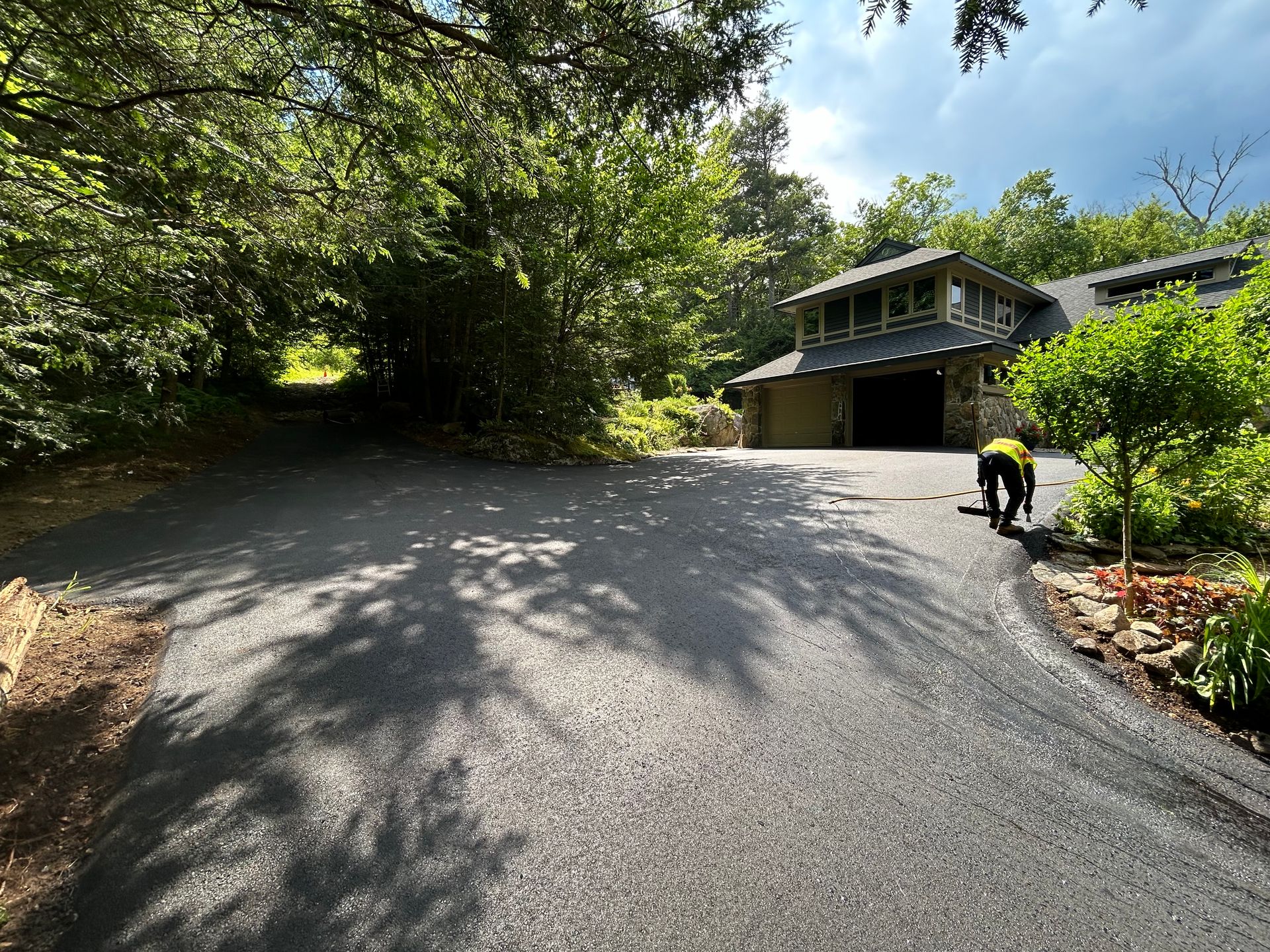 A person in a yellow vest walks on a newly paved asphalt driveway in front of a house surrounded by trees.