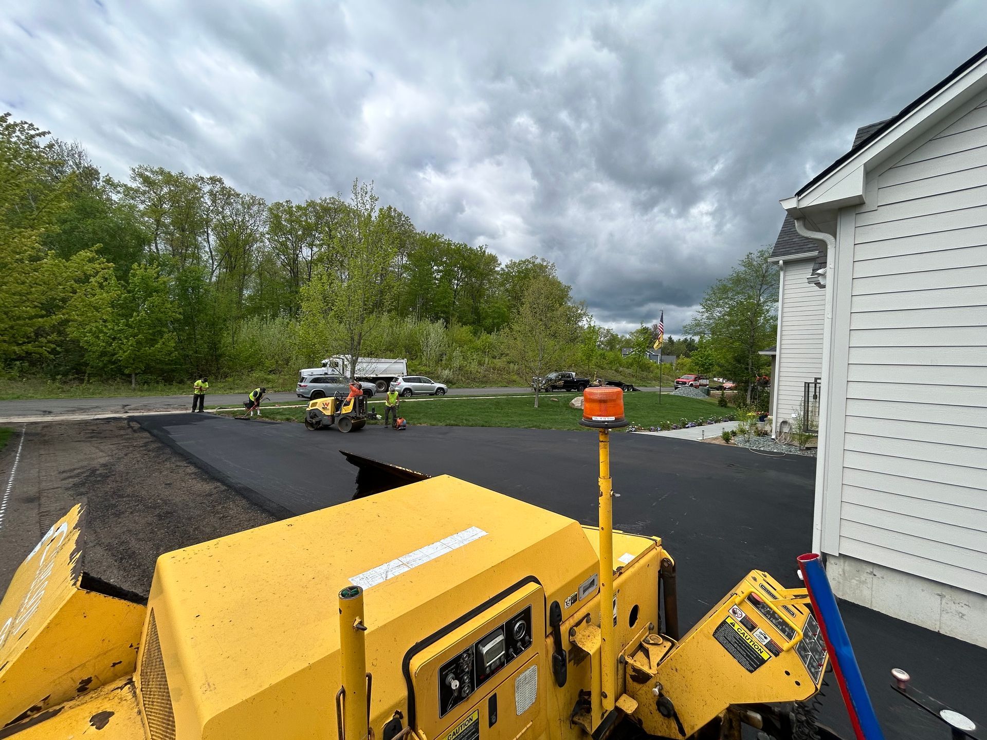 A yellow asphalt paver sits in the foreground as workers pave a driveway next to a white house.