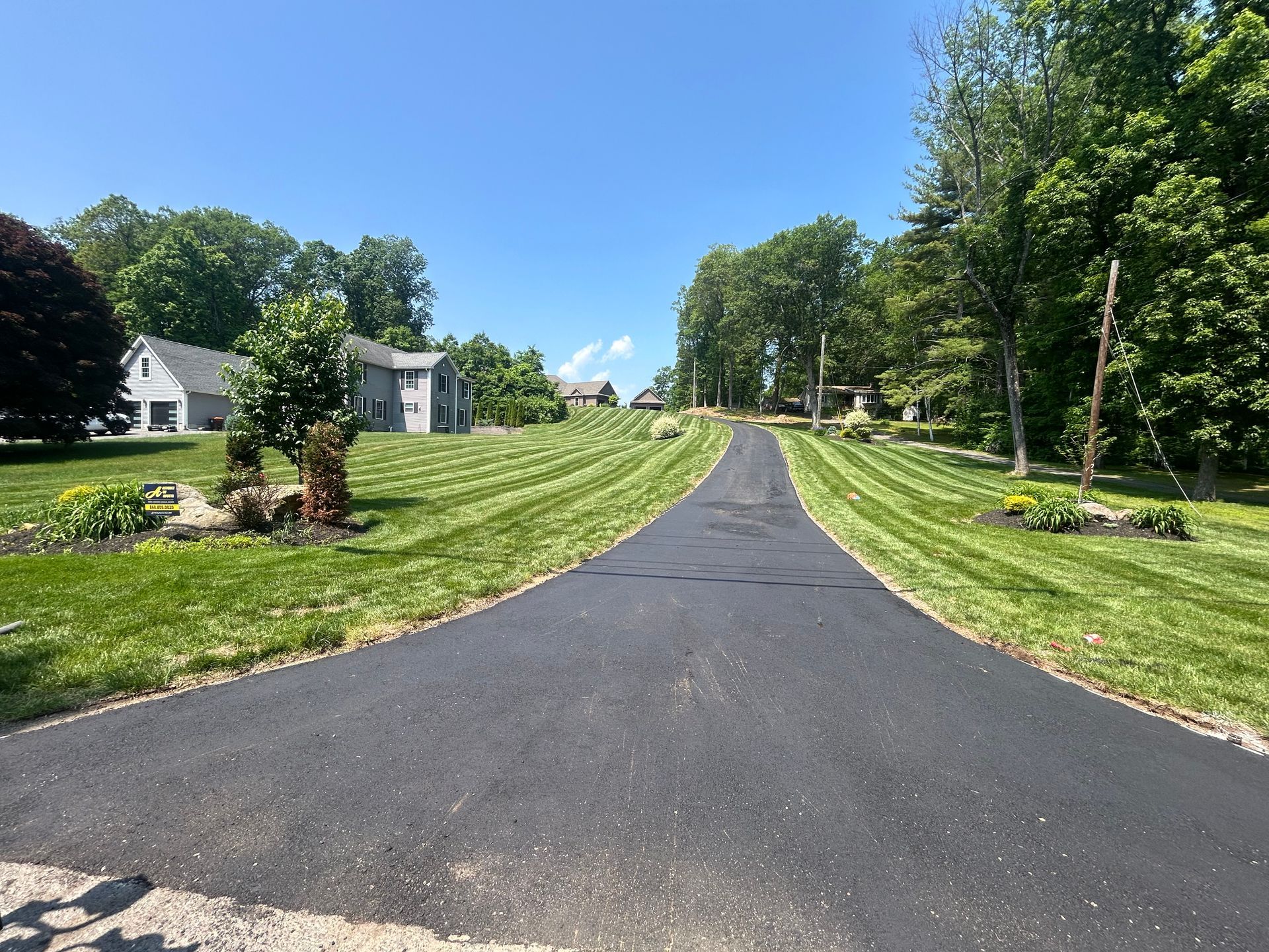 A paved driveway leads up a hill through a lush green yard towards houses under a clear blue sky.