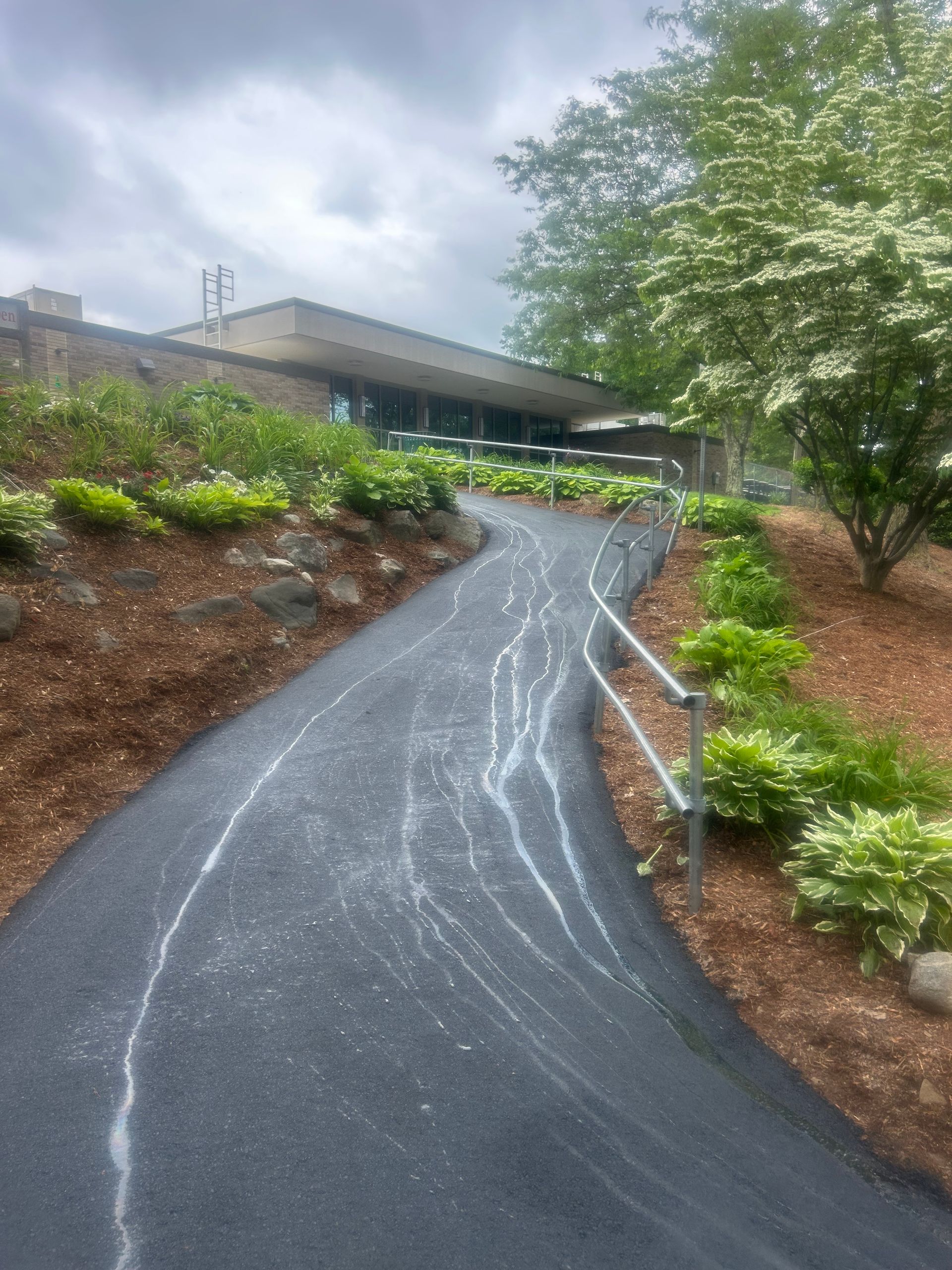 A paved, curved ramp with a metal handrail leads upward toward a modern building entrance surrounded by green landscaping.