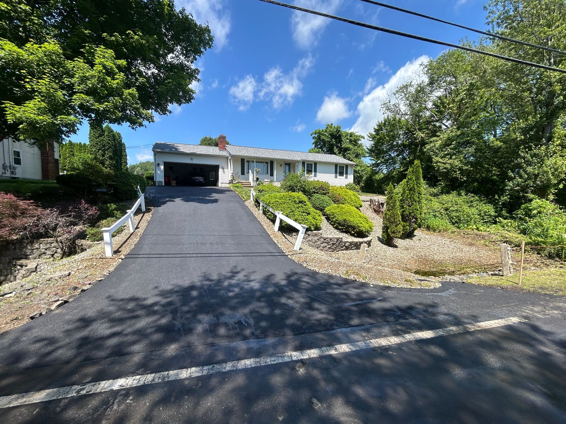 A single-story house with a white exterior and a dark garage at the top of a steep, freshly paved asphalt driveway.