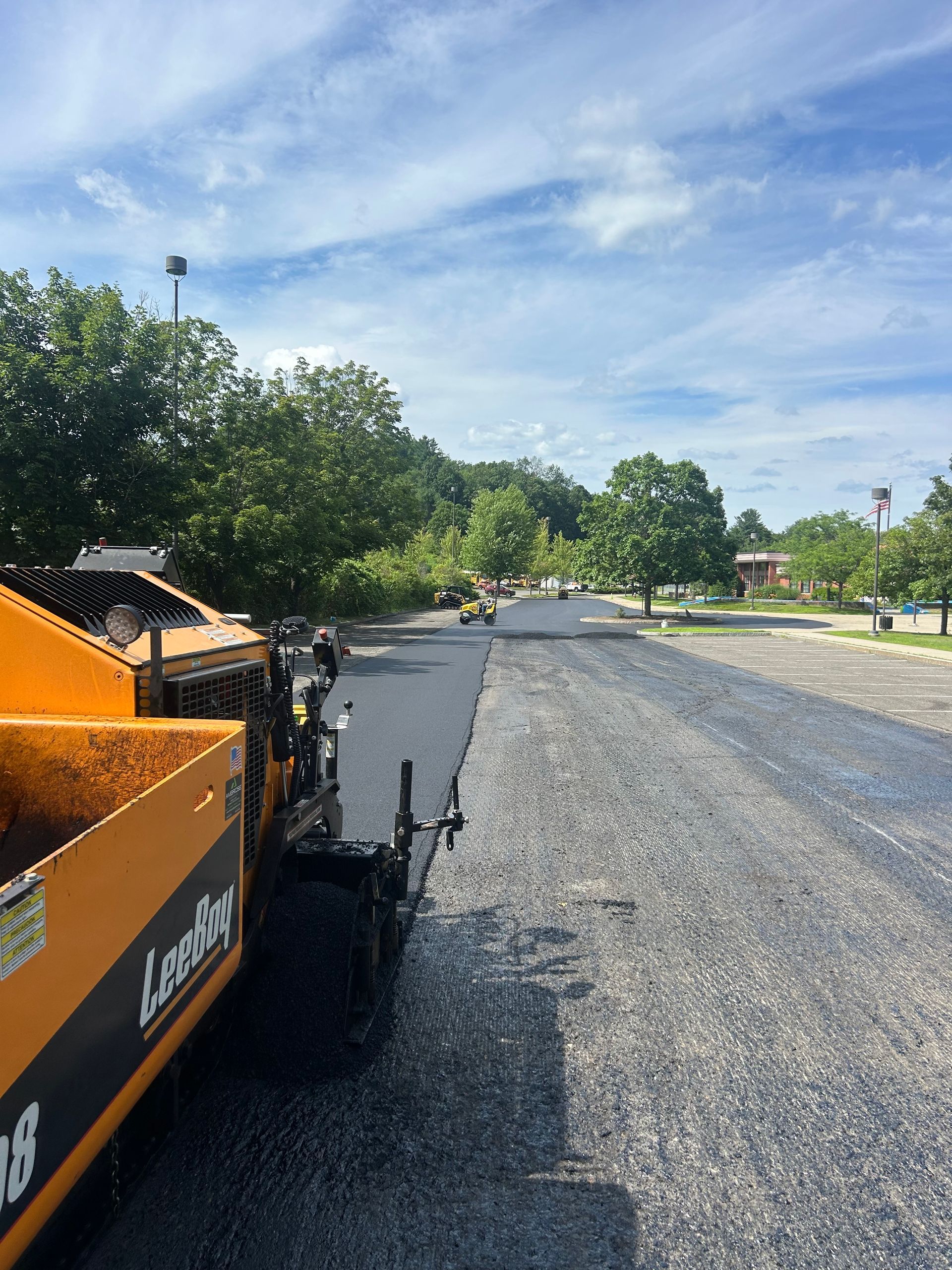 A yellow asphalt paver sits on a sunny day next to a newly paved dark road and an adjacent section of rough gravel.