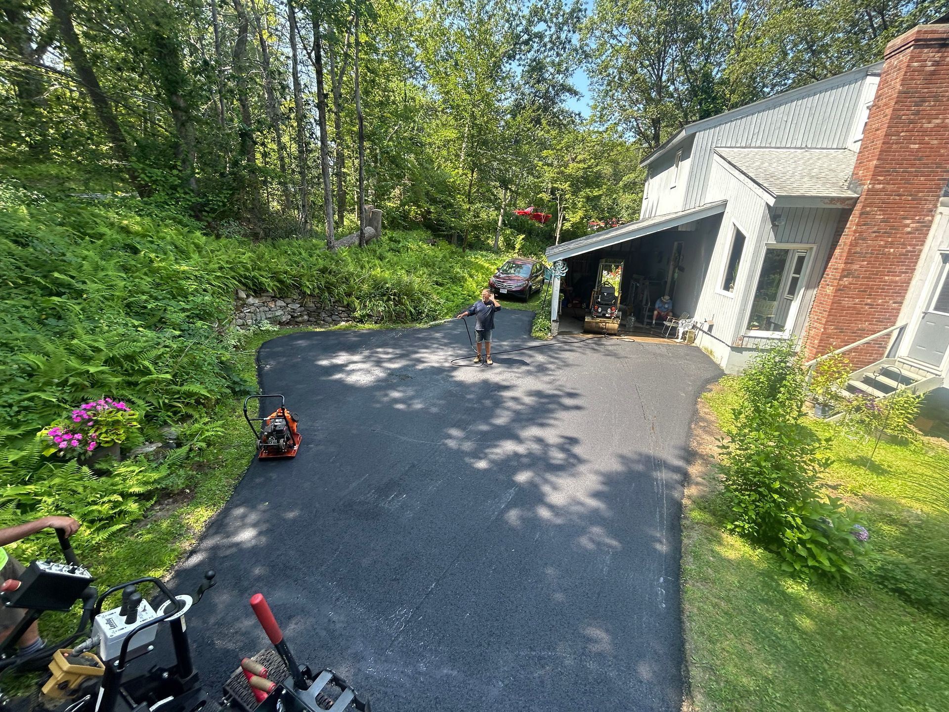 A freshly paved black asphalt driveway leading to a house with a brick chimney, surrounded by green trees and foliage.