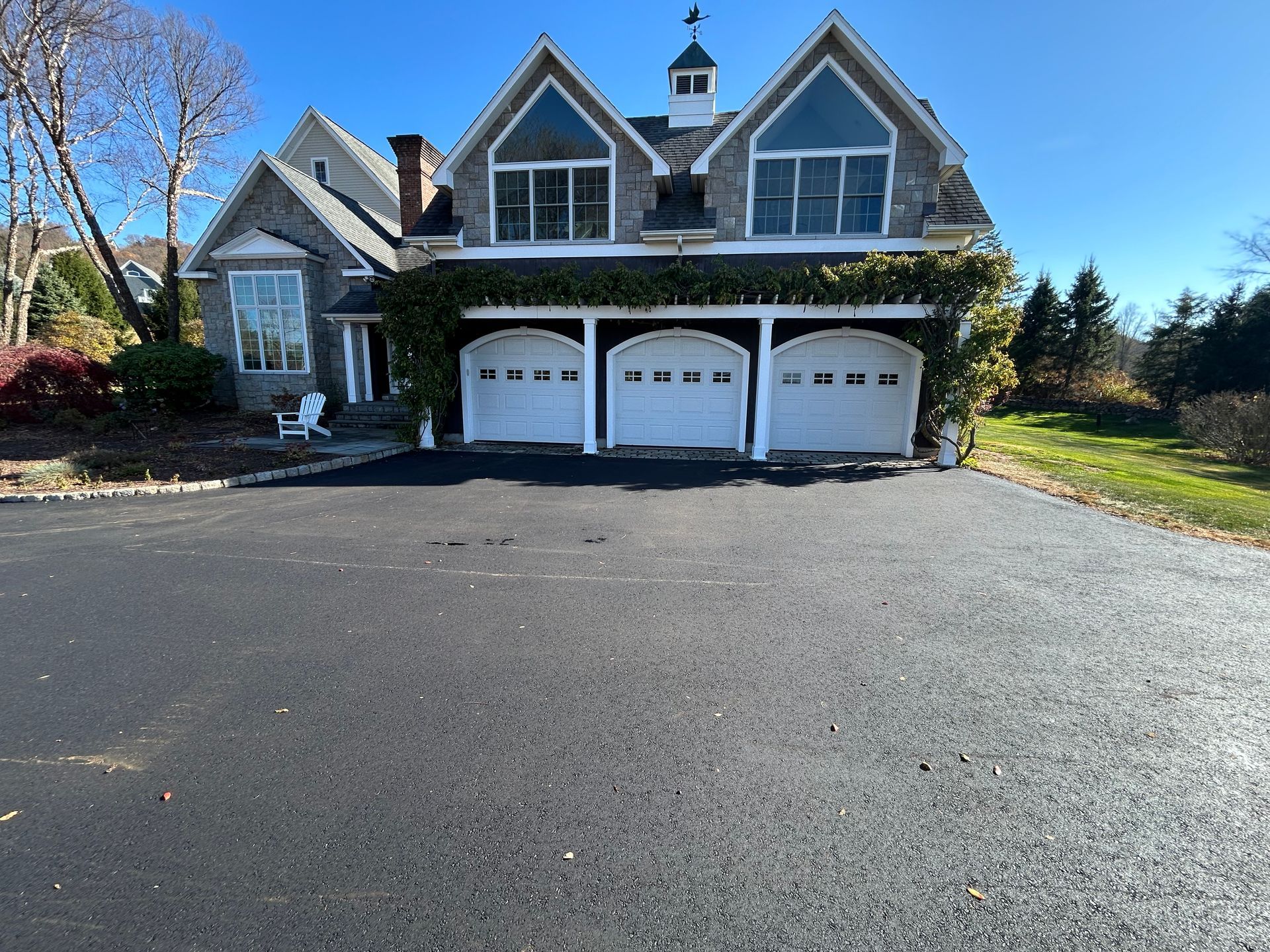 A large, multi-story stone house with a three-car garage and a wide asphalt driveway under a clear blue sky.