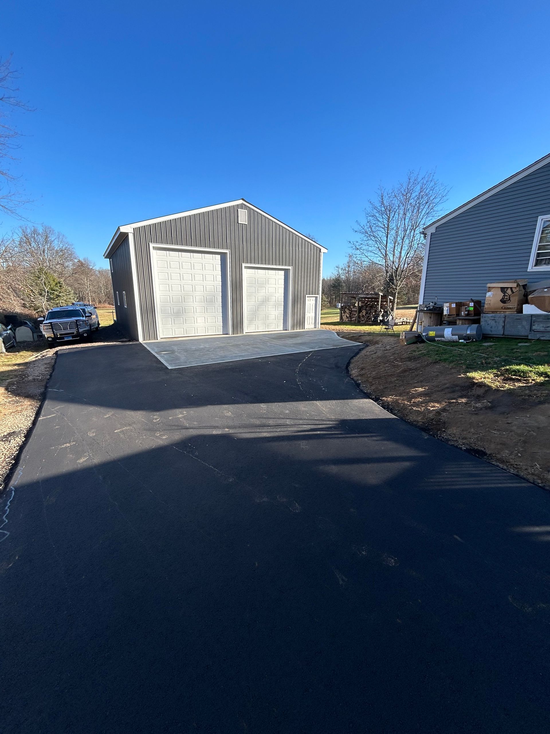 A metal garage with two doors sits at the end of a newly paved black asphalt driveway next to a gray house.