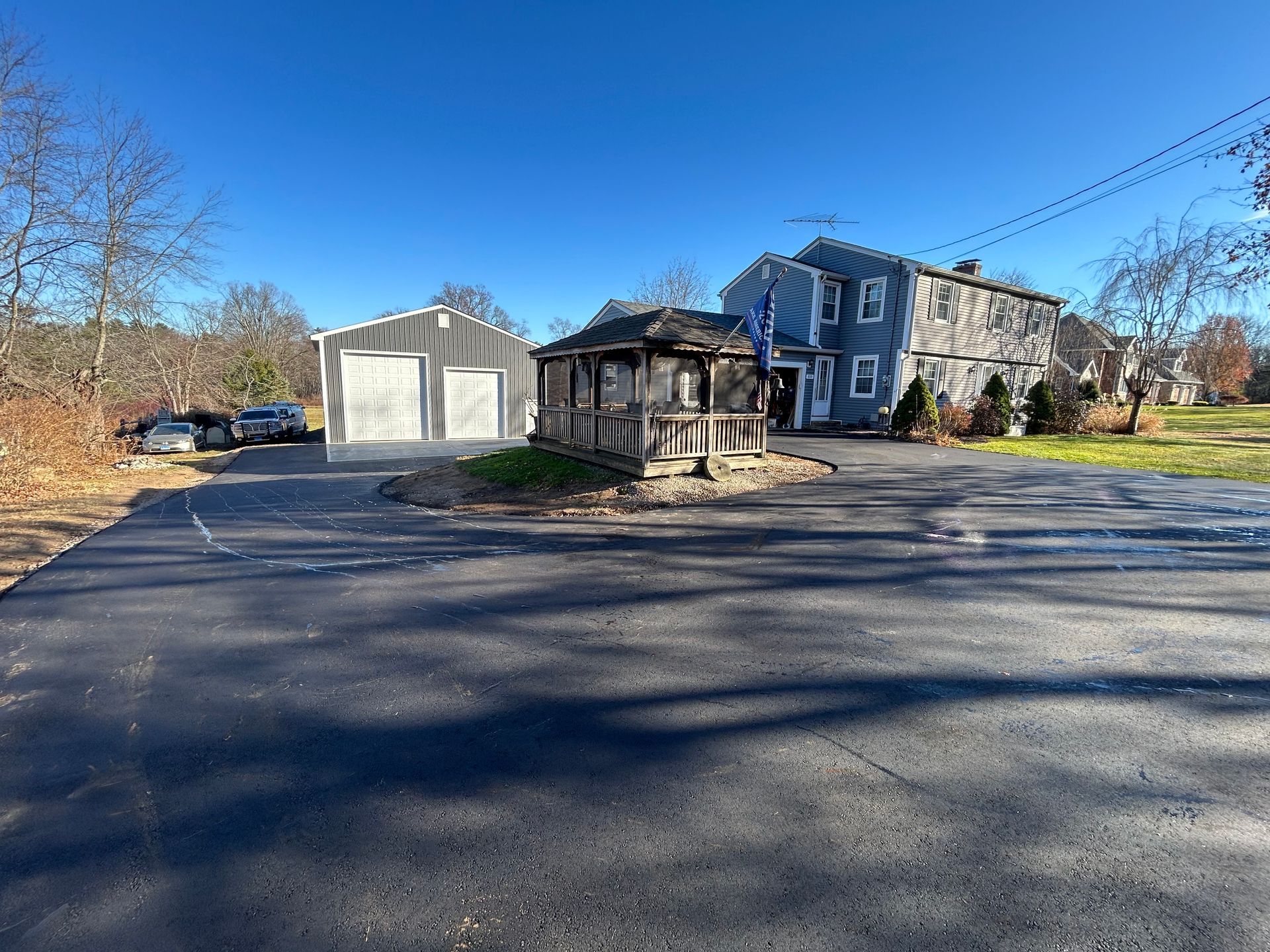 A paved driveway leads to a grey house and a detached metal garage under a clear blue sky on a sunny day.