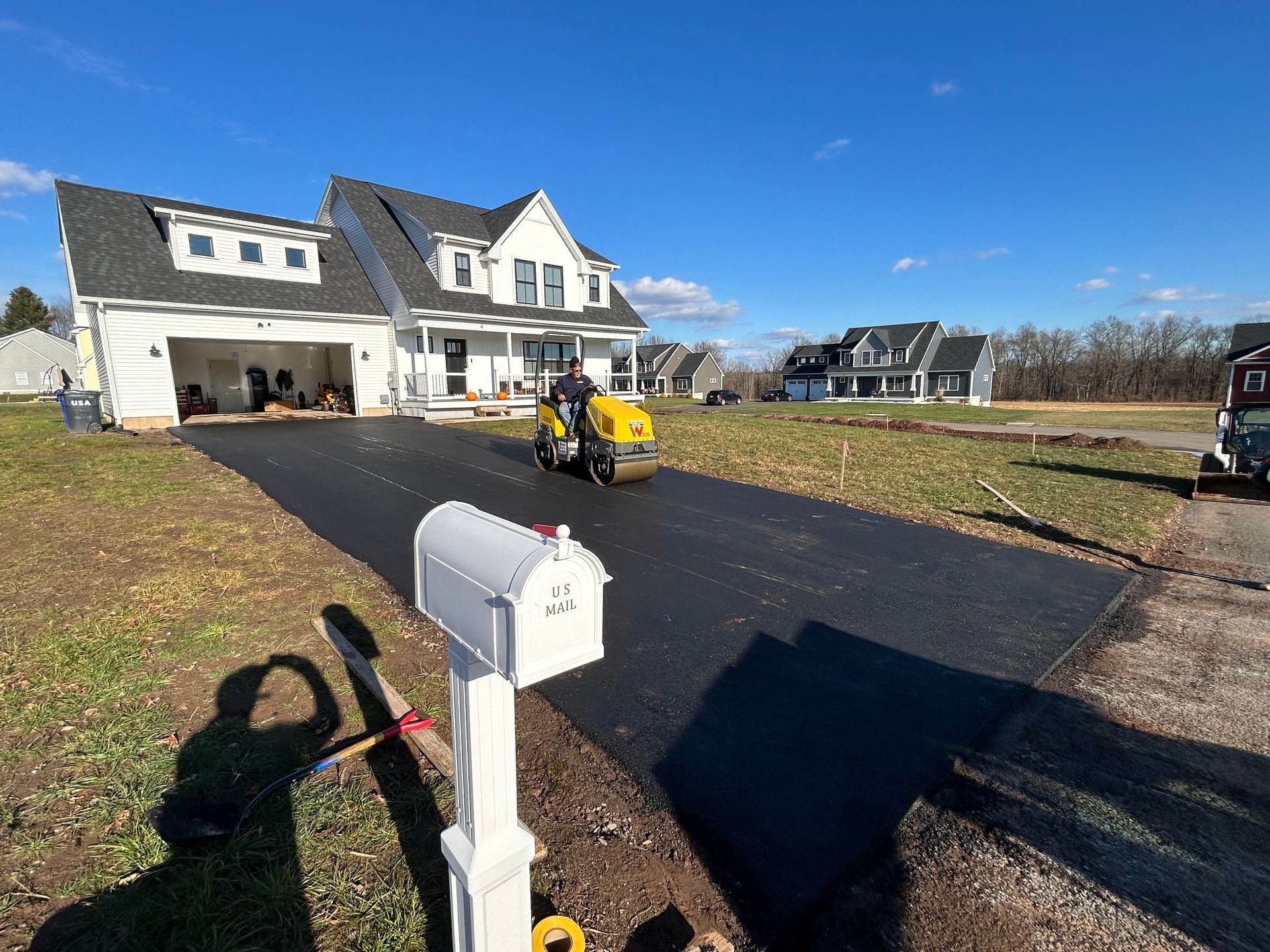 A person drives a yellow steamroller over a newly paved asphalt driveway in front of a white two-story house.