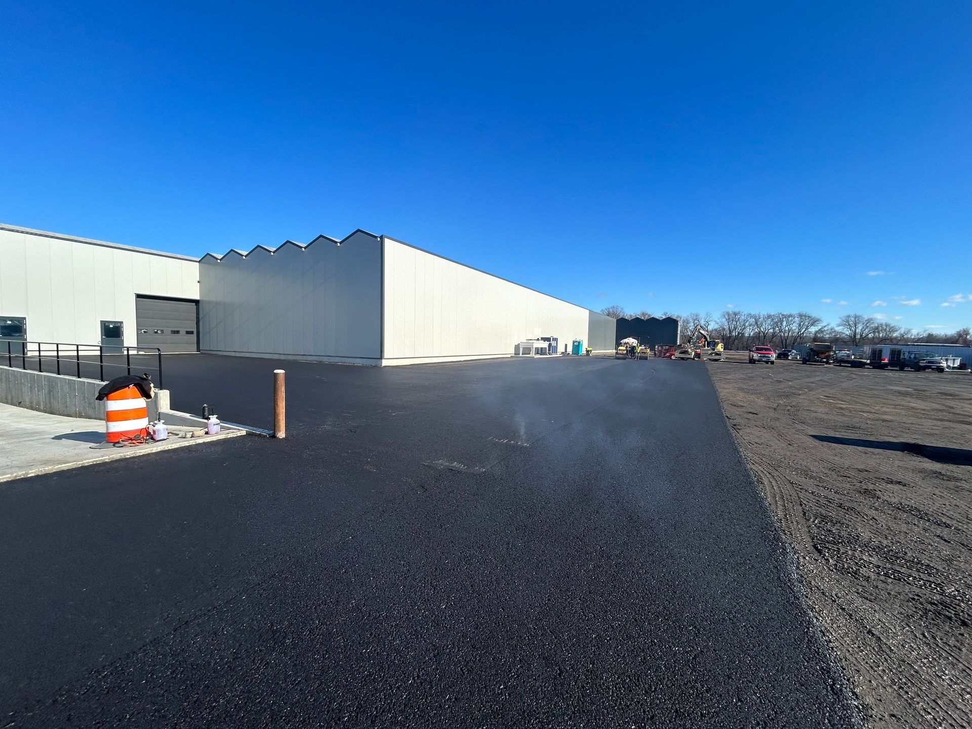 Freshly paved asphalt driveway leading to a large, white industrial warehouse building under a clear blue sky.