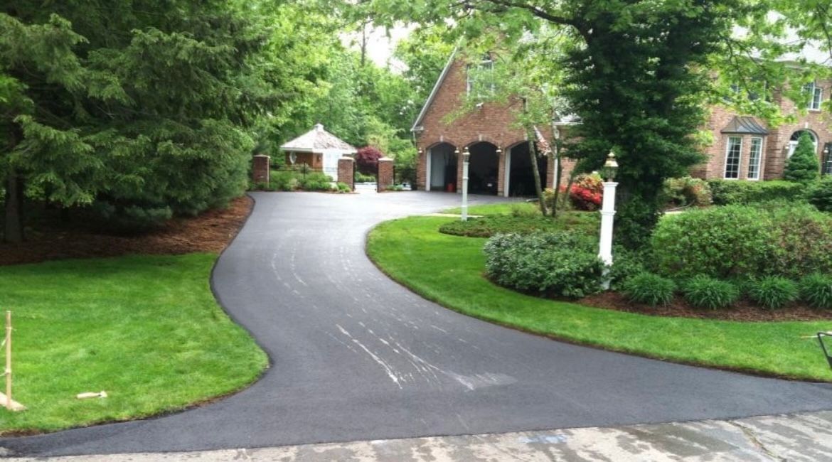 A freshly paved, curved asphalt driveway leads toward a brick house with a multi-car garage on a sunny, grassy property.