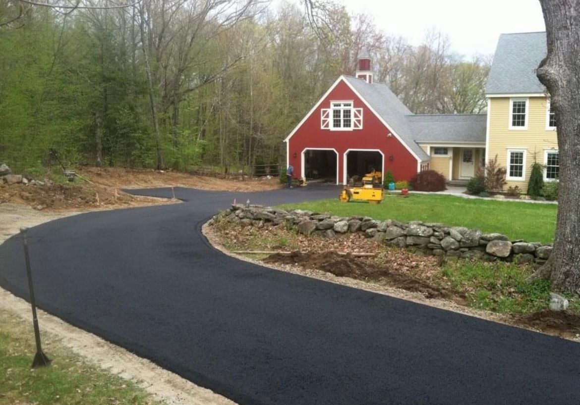 A newly paved, curved asphalt driveway leads toward a red garage attached to a yellow house surrounded by trees.