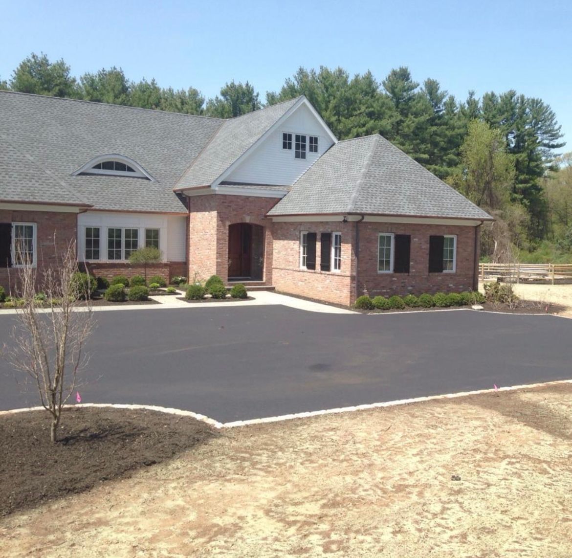 A large, single-story brick house with a gray shingled roof, a paved driveway, and a partially landscaped yard.