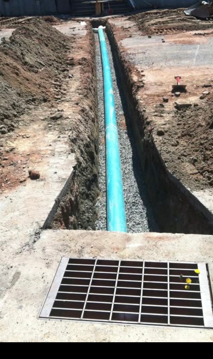 A long blue pipe runs through a gravel-lined trench in the ground, leading toward a metal grate in the foreground.