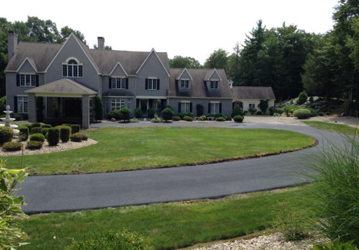A large, grey, multi-gabled house with a curved asphalt driveway, front lawn, and trees under a clear sky.