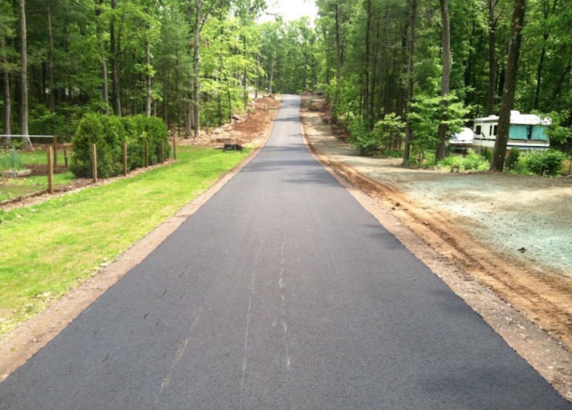 A newly paved asphalt driveway stretches through a wooded area, bordered by a green lawn on the left and trees on the right.