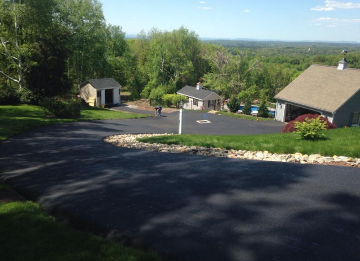 A paved driveway winds past a small shed and a house toward a garage on a sunny, tree-lined property with a distant view.