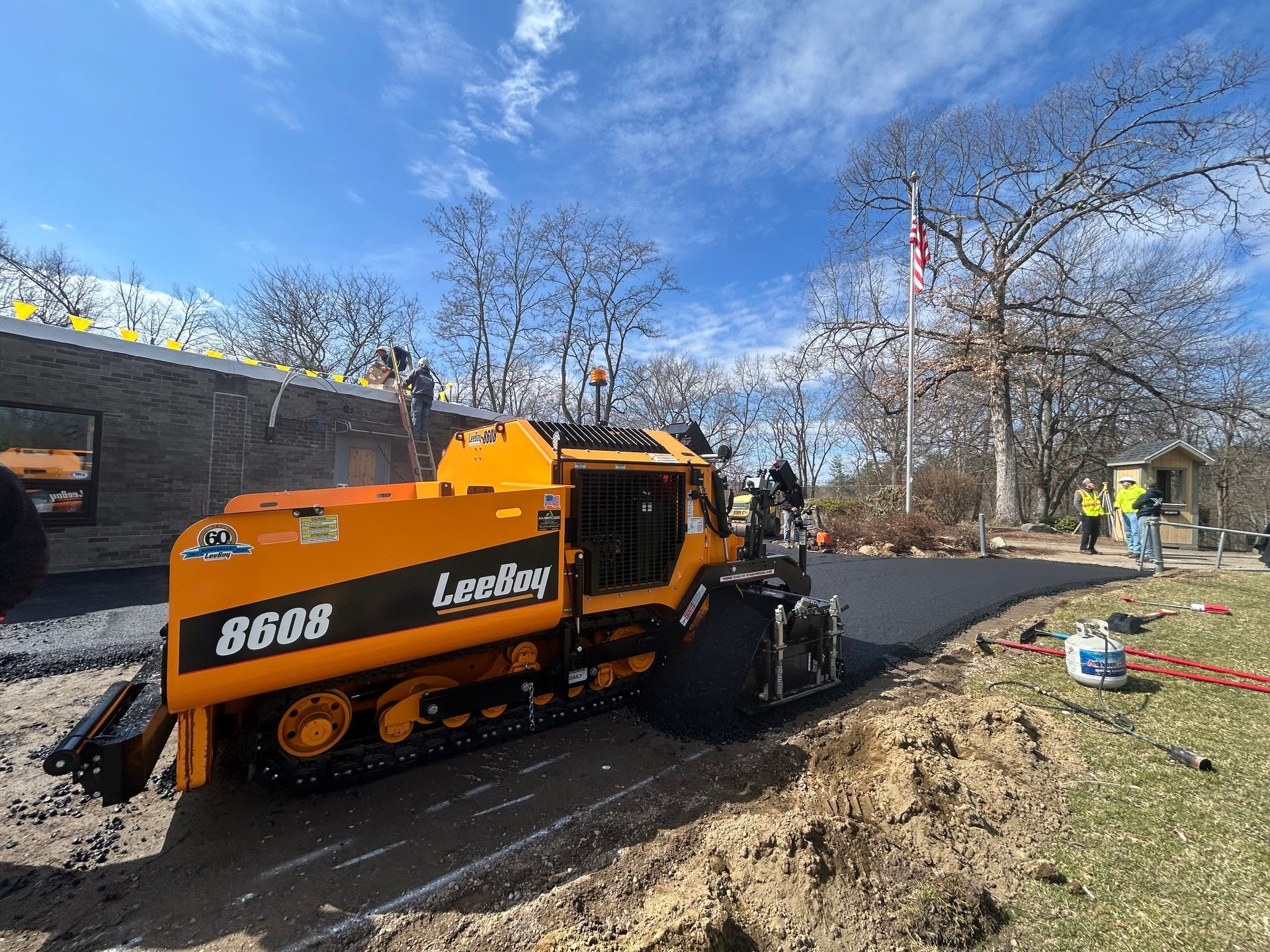 A yellow LeeBoy 8688 asphalt paver operates on a paved driveway next to a building and trees under a clear blue sky.