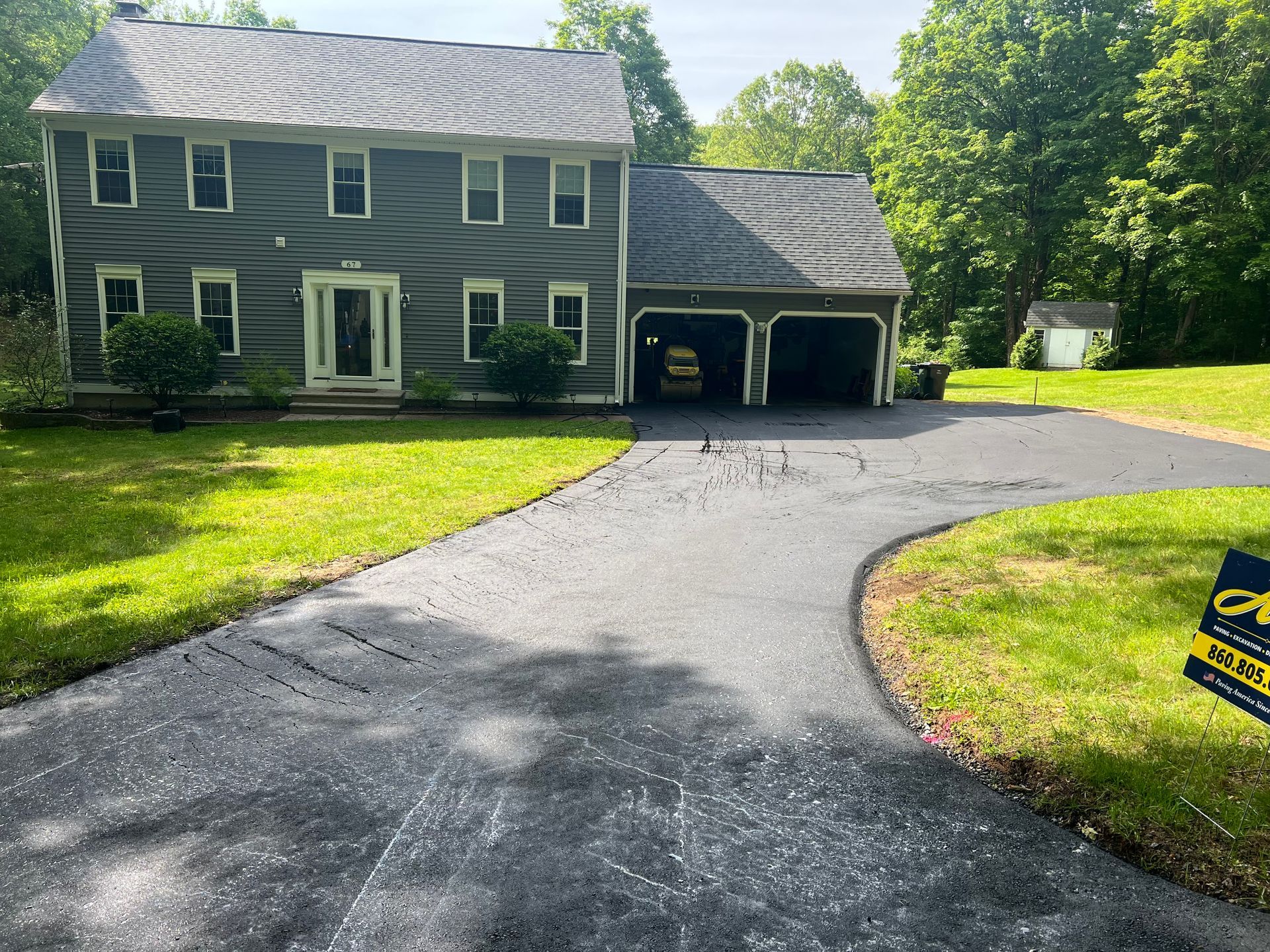A gray two-story house with a two-car garage and a newly paved driveway, surrounded by a green lawn and trees.