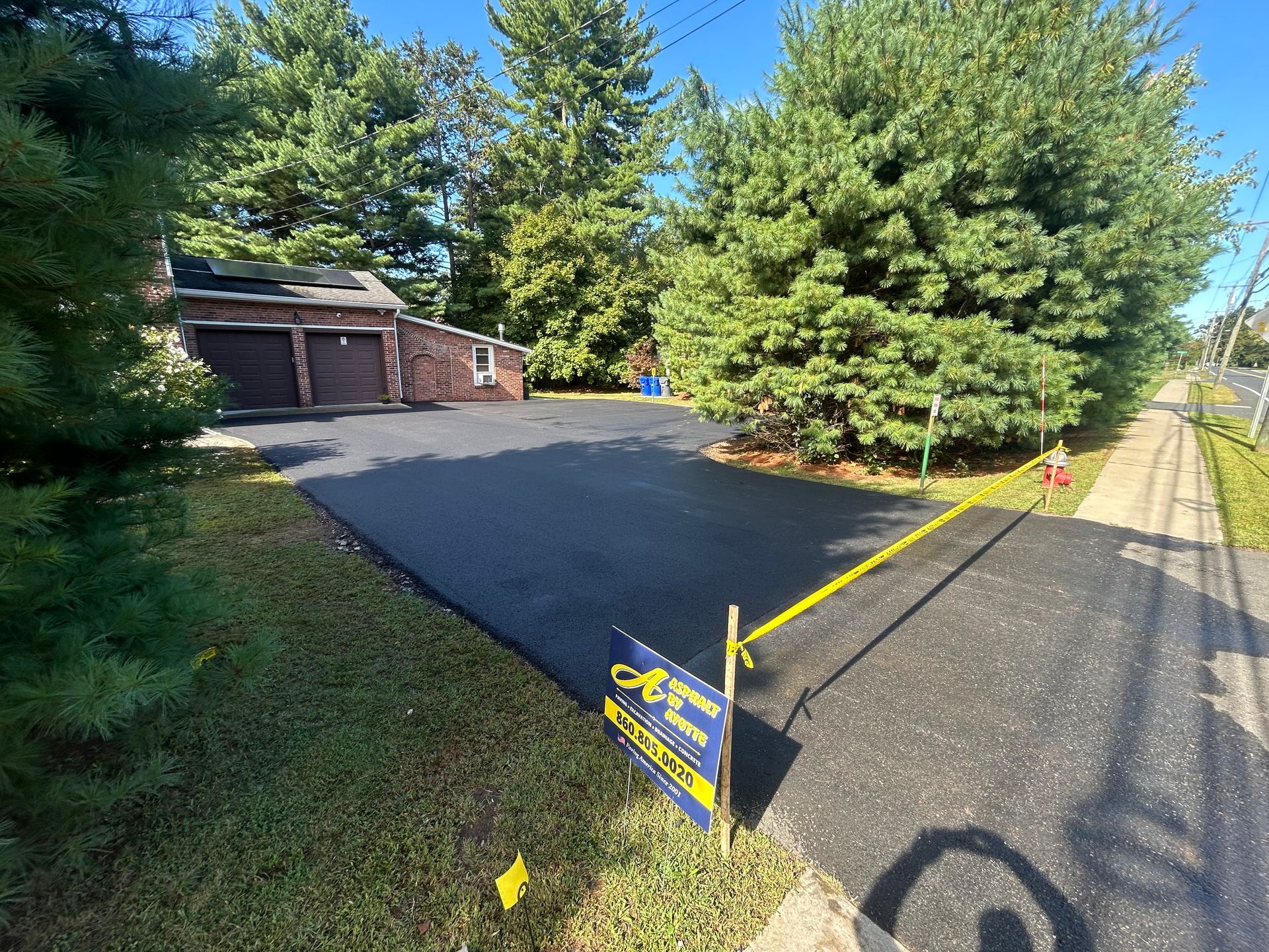 A newly paved dark asphalt driveway leads to a brick house garage, with a business sign marked off by yellow caution tape.