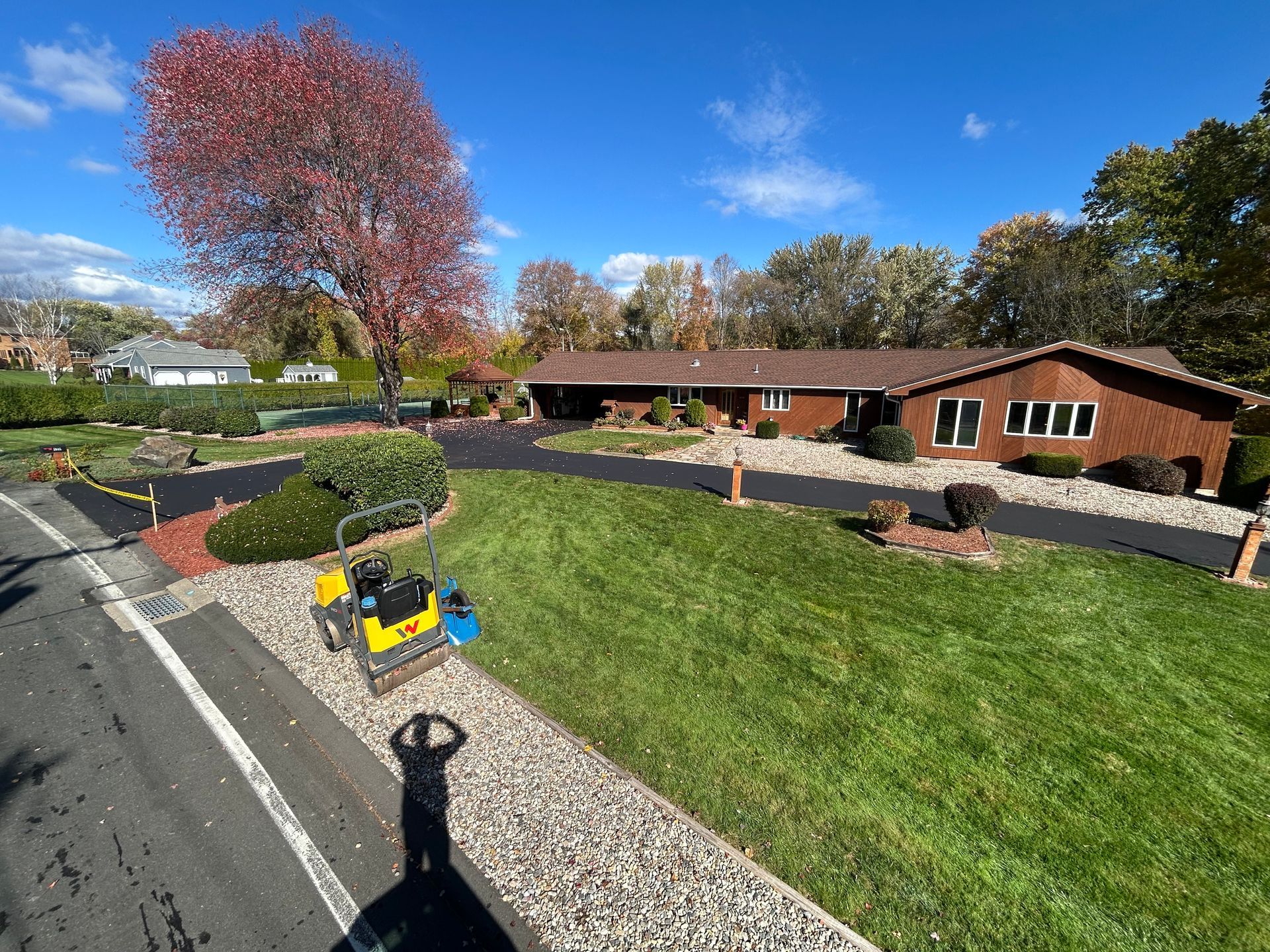 A construction roller sits on gravel in front of a newly paved driveway leading to a suburban brick home on a sunny day.
