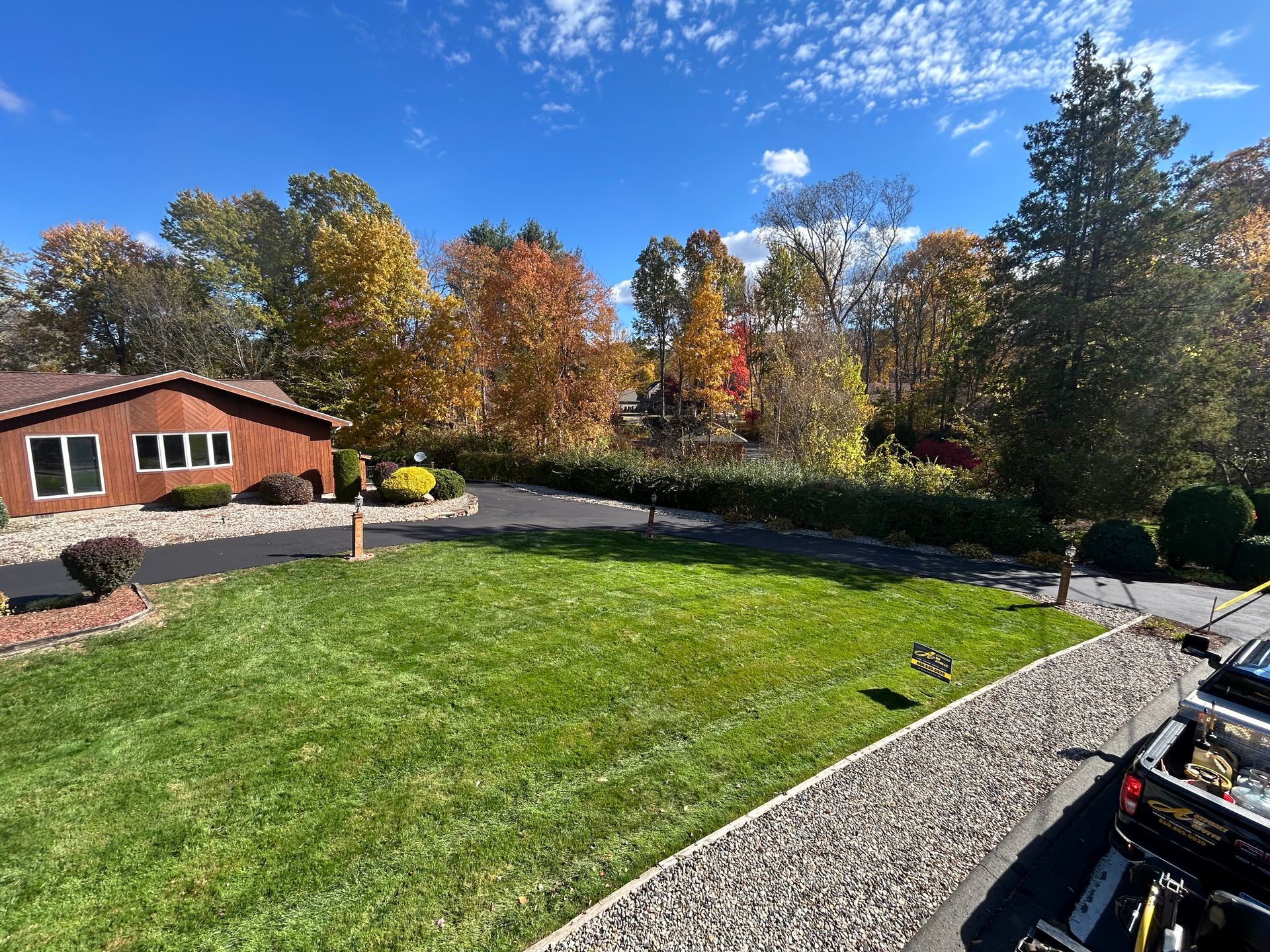 A sunny suburban yard with a mown lawn, a gravel border, and a wooden house surrounded by colorful autumn trees.