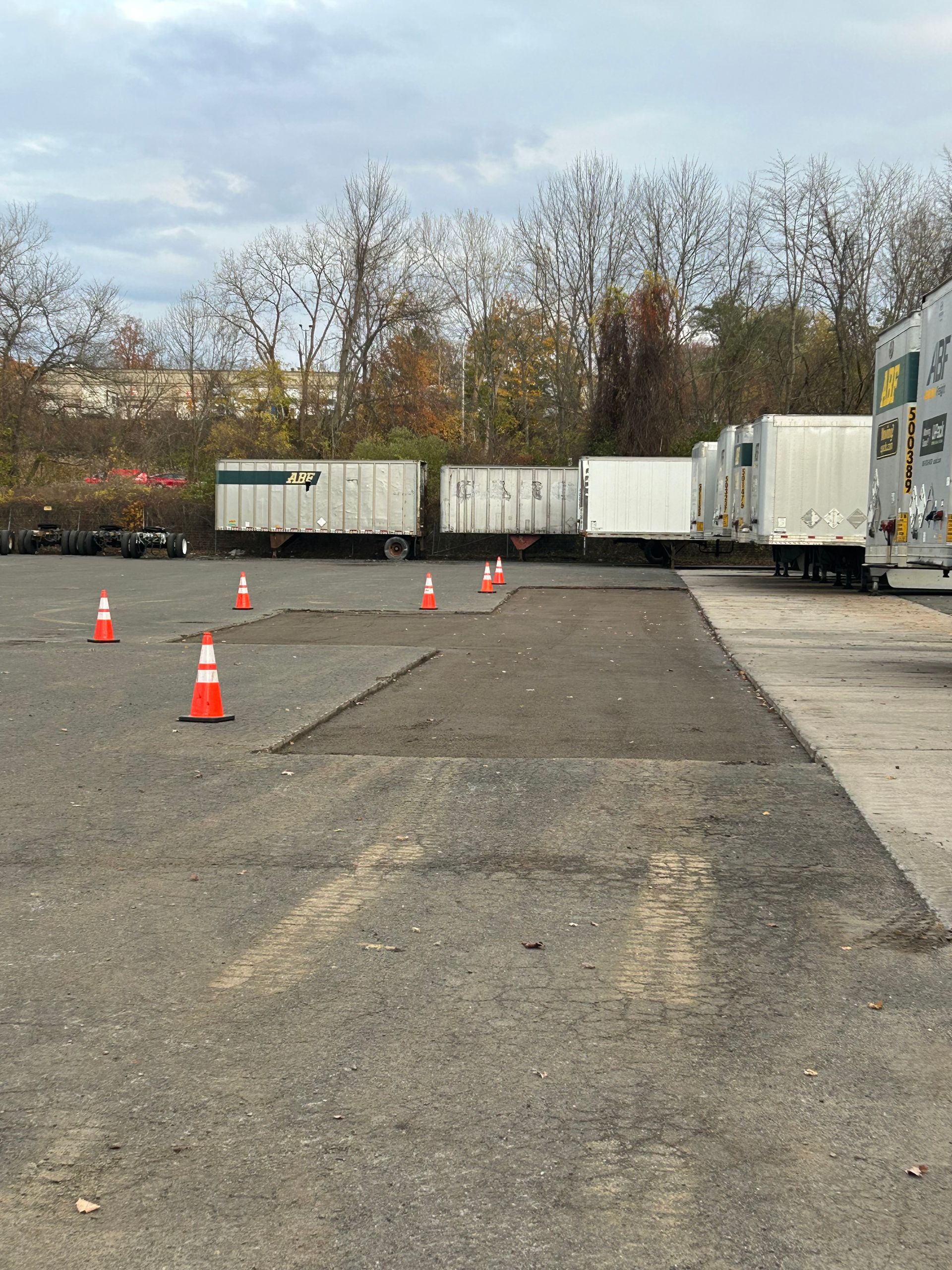 A row of parked white semi-trailers in a gravel lot with orange safety cones demarcating a patch of fresh pavement.