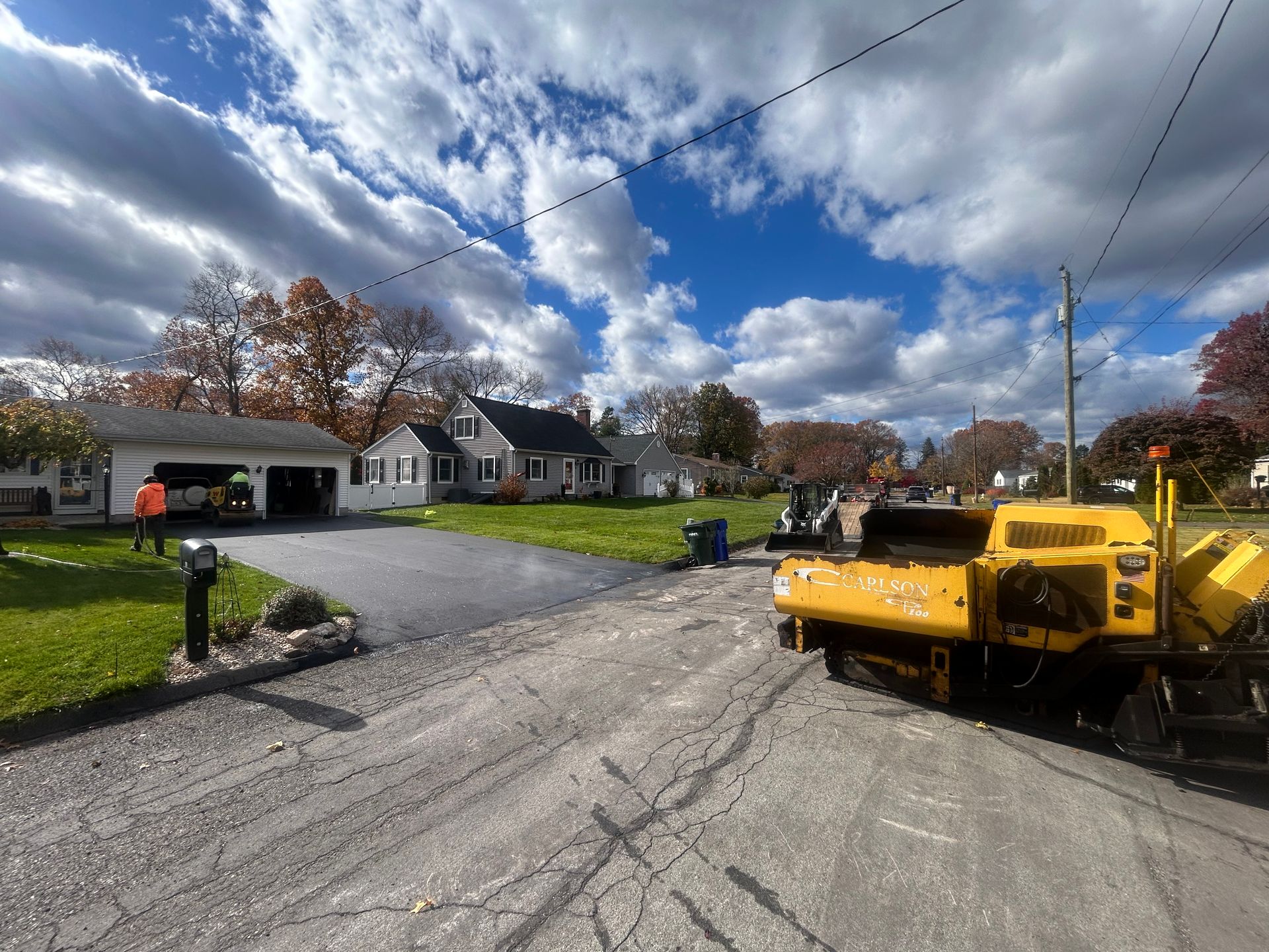 Construction workers pave a driveway in front of a suburban house, with a yellow paving machine parked on the road.