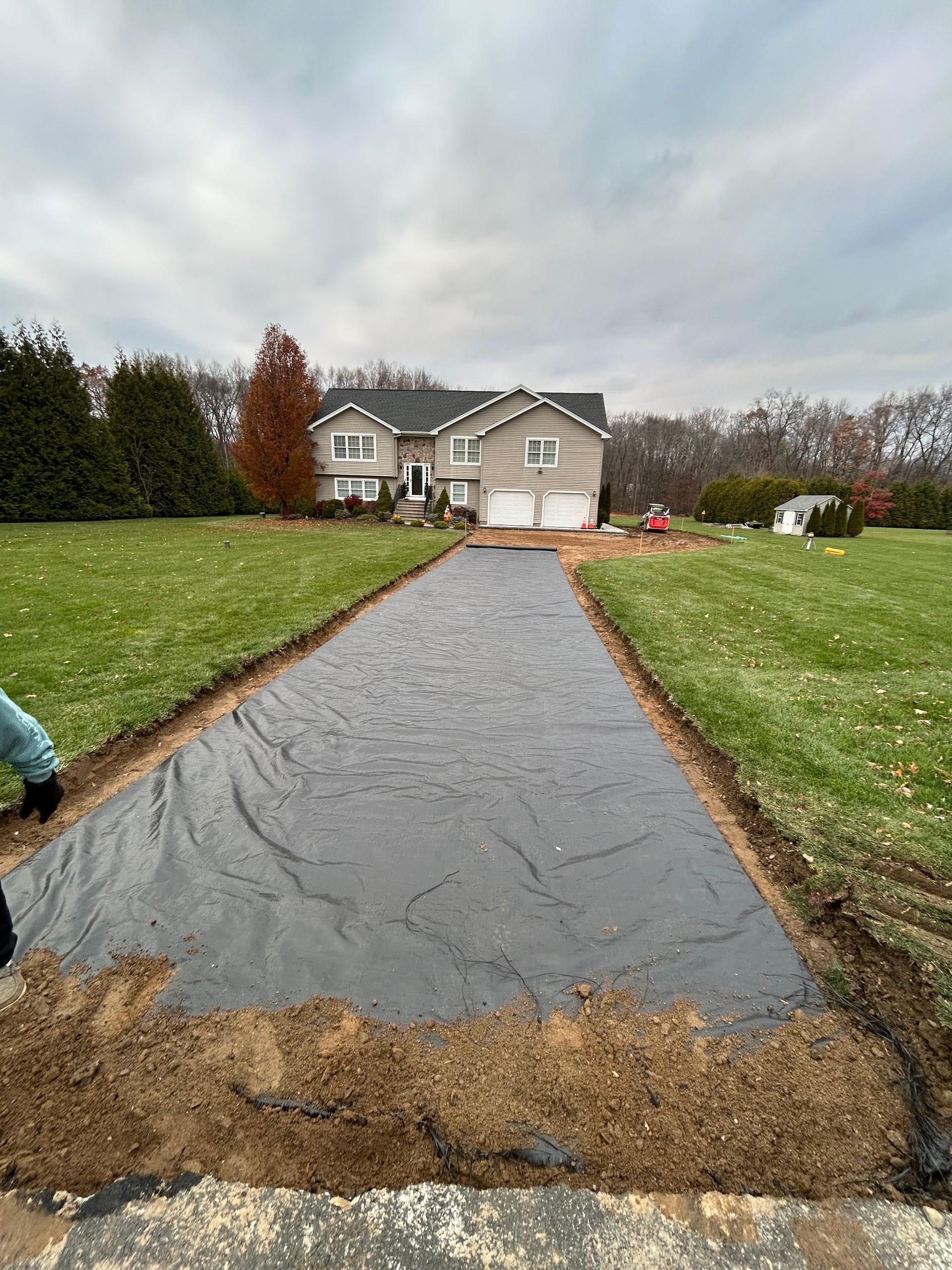 A long, excavated driveway covered with black landscape fabric leading to a suburban house under a cloudy sky.