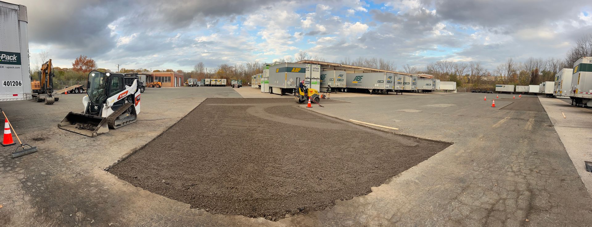 A panoramic view of an outdoor construction site with gravel, a skid steer, and multiple portable buildings in the distance.
