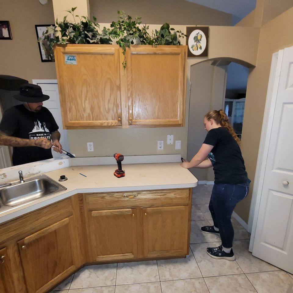 Two people removing a kitchen countertop and cabinets; one uses a tool, the other supports.