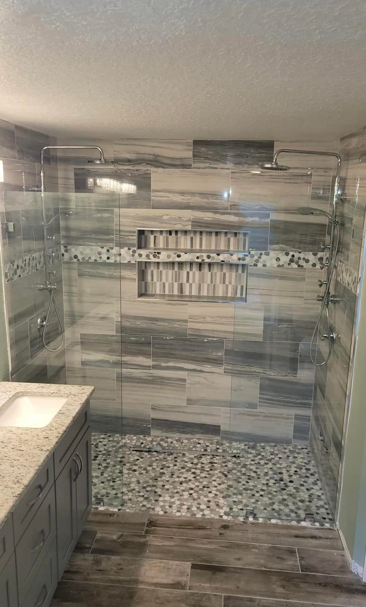 Modern shower with gray and white tile, pebble floor, glass walls, and built-in shelves.