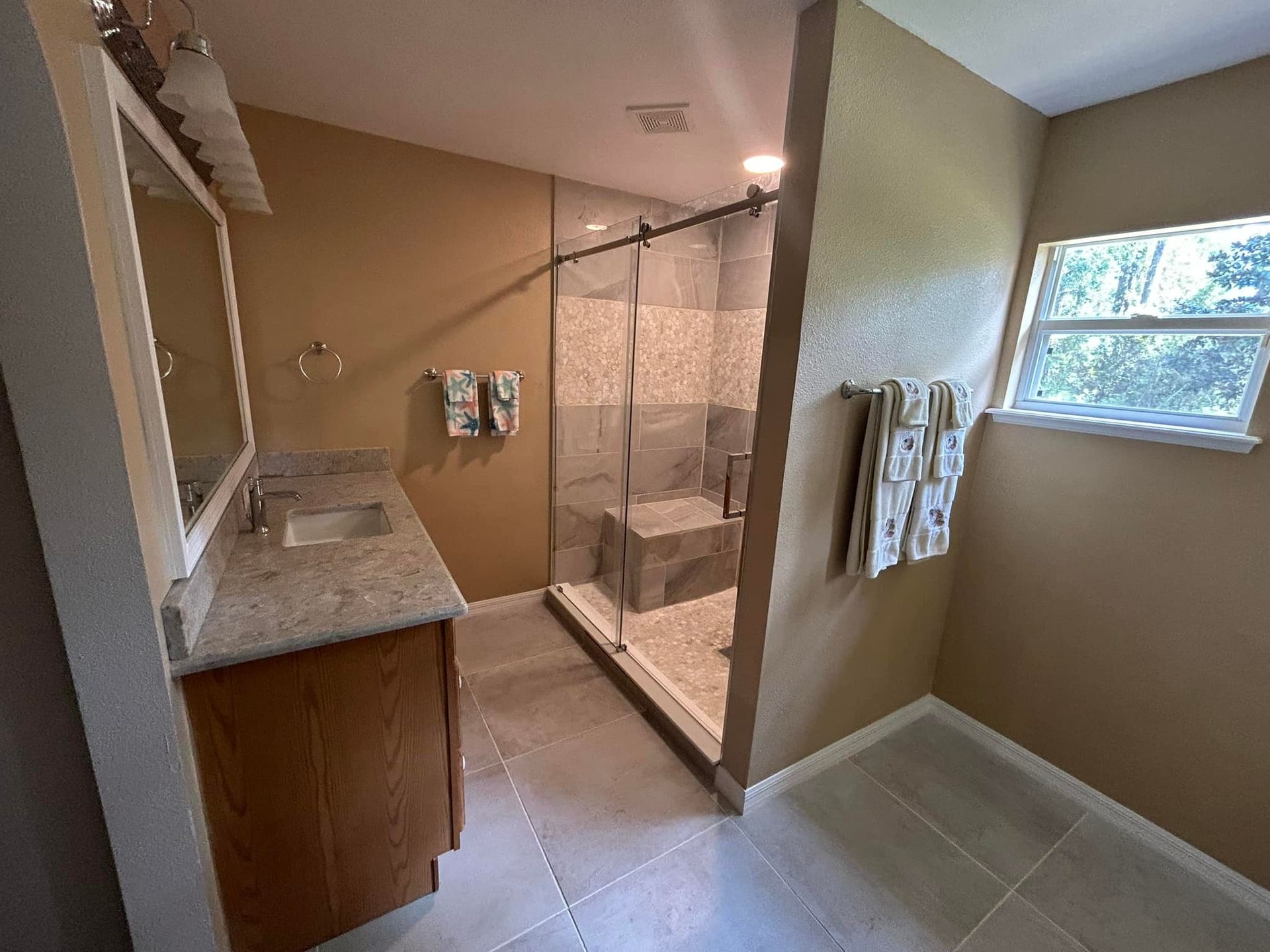 Bathroom with a walk-in shower. Beige walls, wood vanity, gray tile floor. Window on the right.