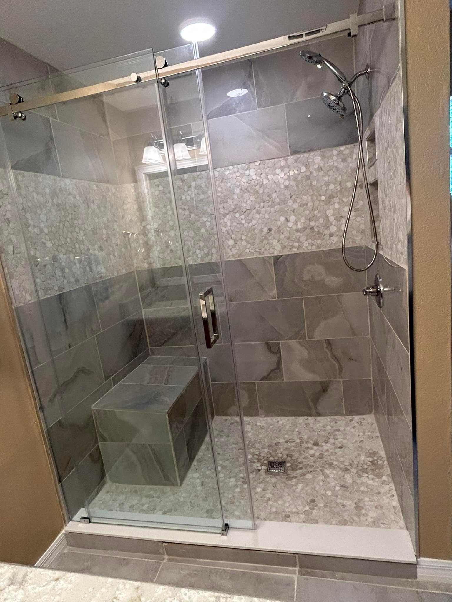 Shower with stone tile walls, glass door, built-in seat, and rain shower head.
