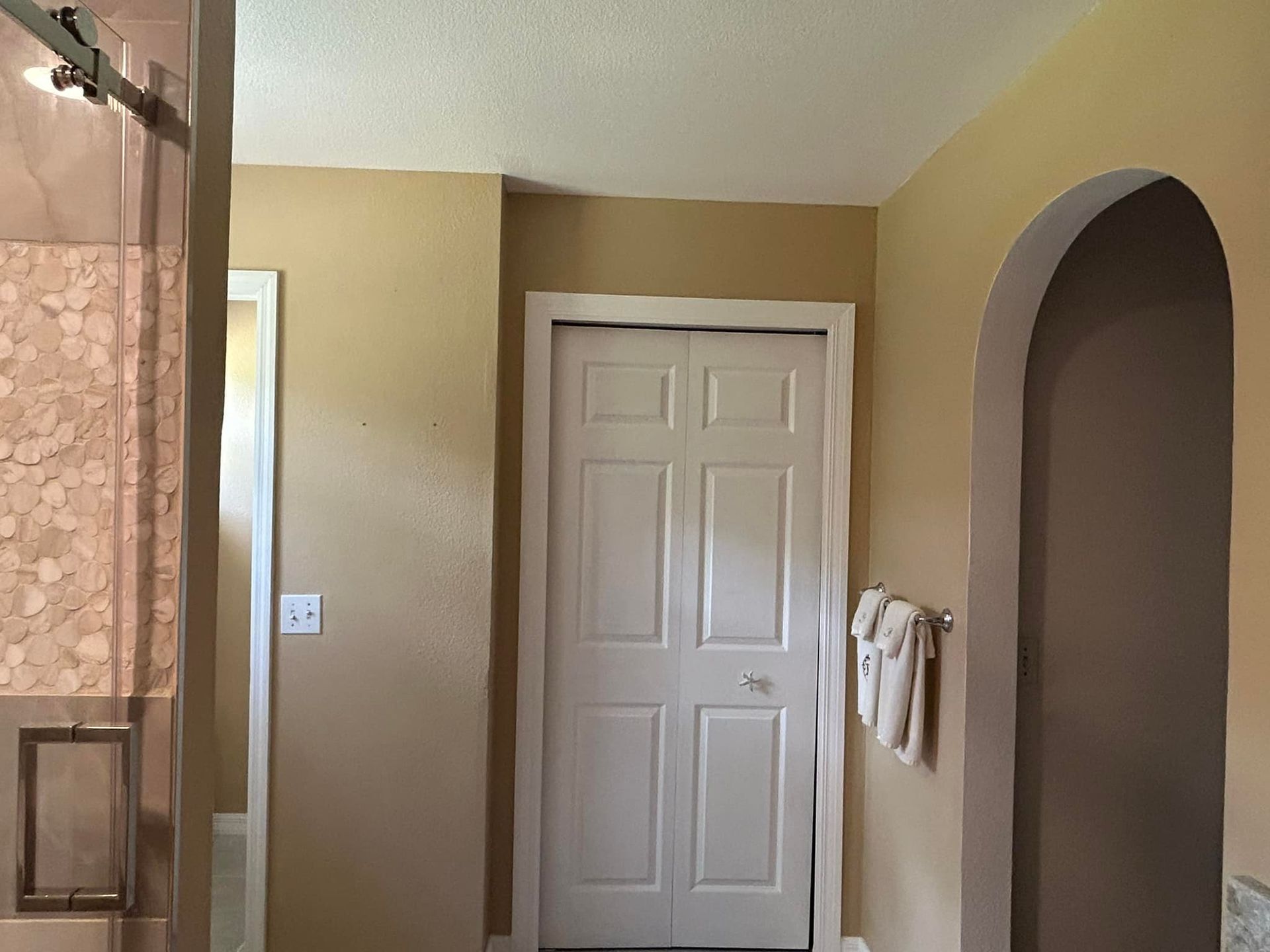 Bathroom interior with shower, closed white door, towel on a hook, and arched doorway. Beige walls and ceiling.