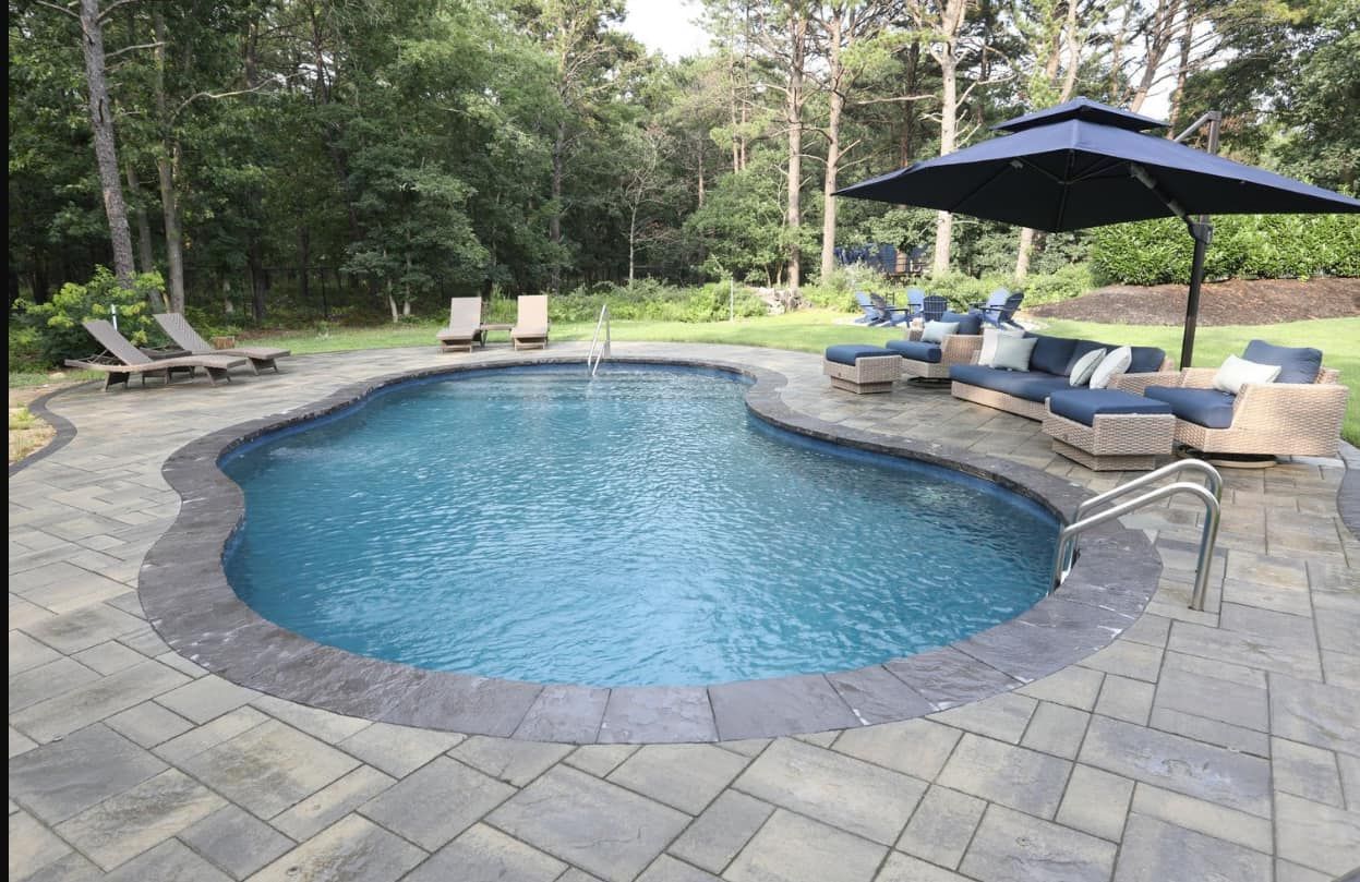 Pool with blue water surrounded by stone patio and lounge furniture, shaded by an umbrella.