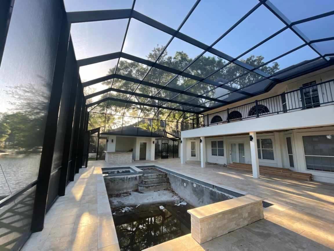 Screened-in pool area with a house. The black screen frames the pool, a hot tub, and a two-story white house on a sunny day.
