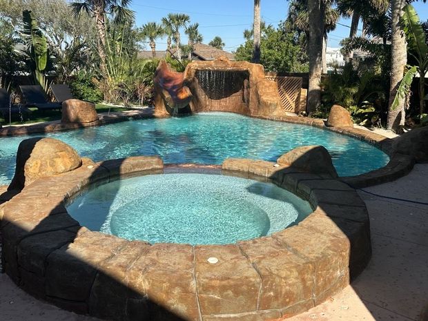 Pool with waterfall feature and attached hot tub, surrounded by stonework, palm trees, and blue sky.