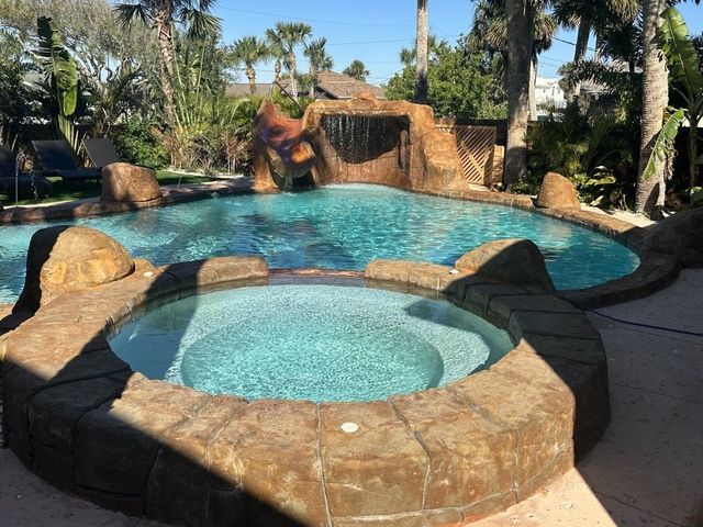 Pool with waterfall feature and attached hot tub, surrounded by stonework, palm trees, and blue sky.