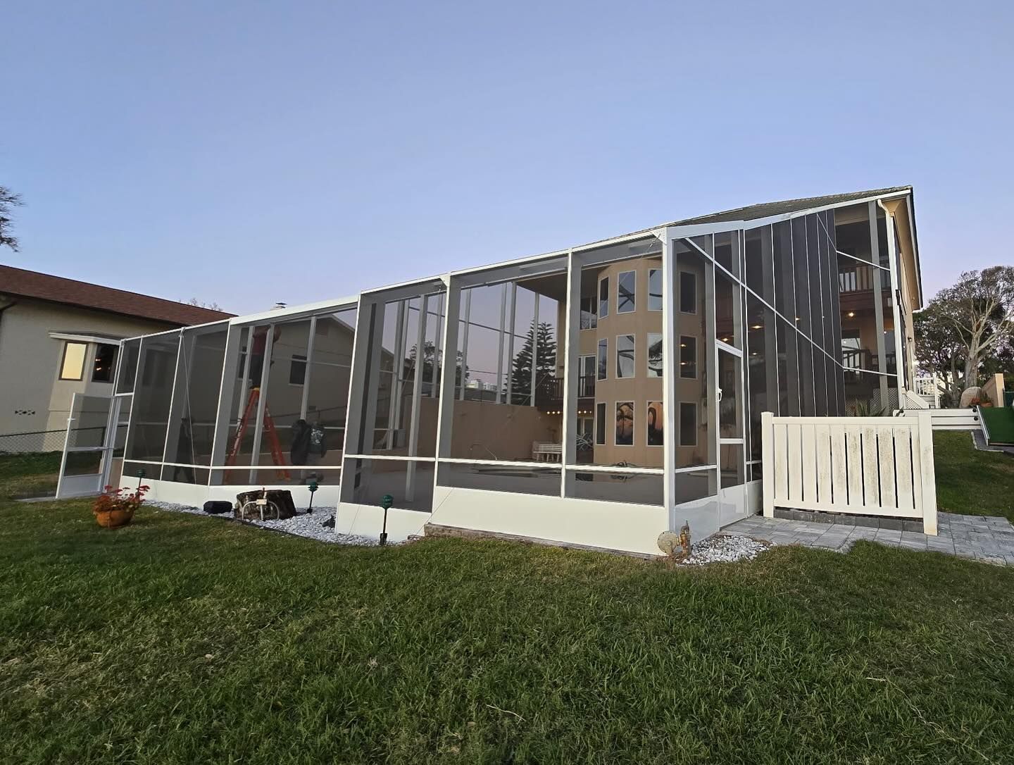 Screened-in patio with white frame, attached to a building, set on a grassy lawn under a pale blue sky.