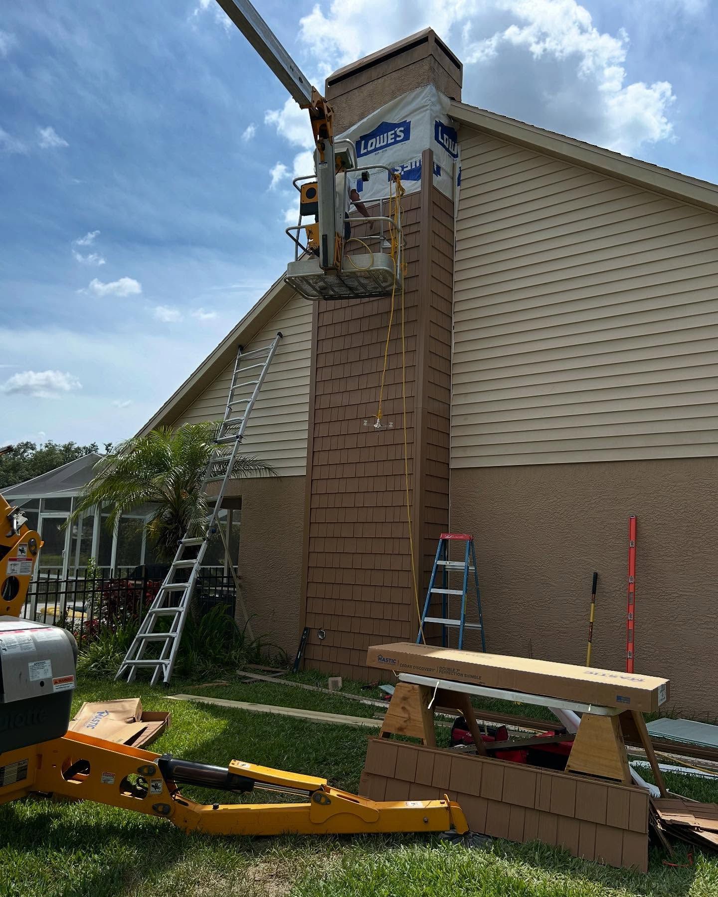 Construction workers repair a chimney on a house with beige siding, using a lift.