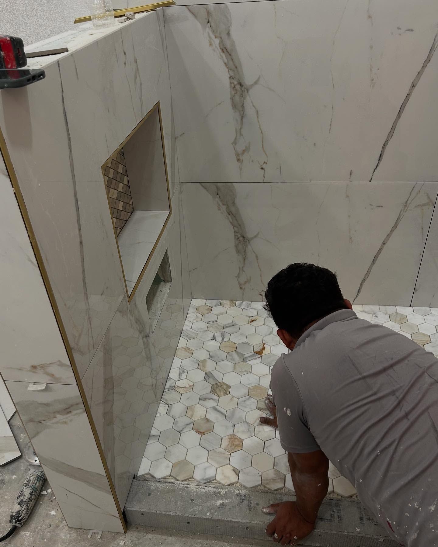 Person tiling a shower floor with mosaic tiles; walls are partially tiled with large marble-look tiles.